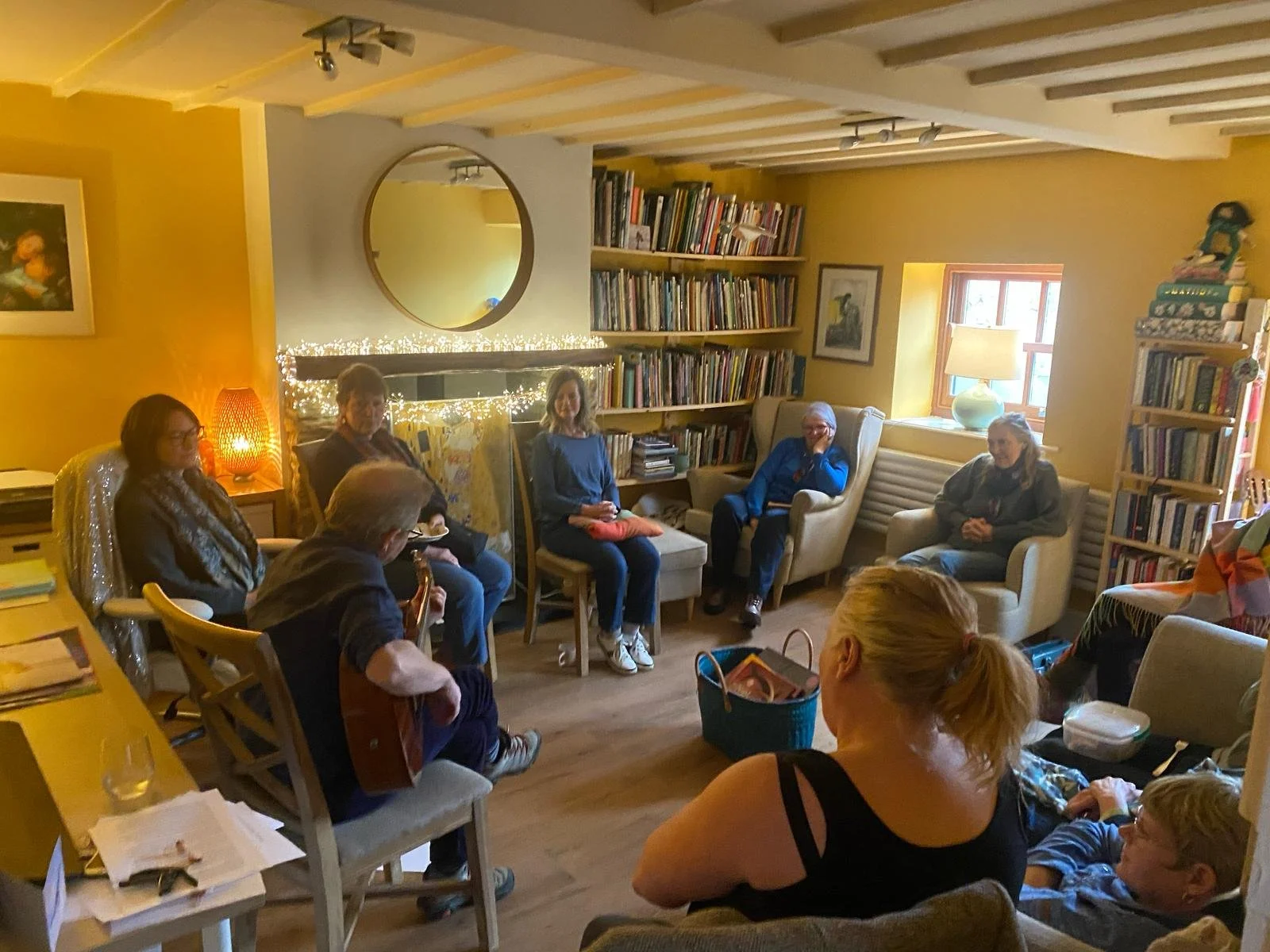 A group of people are gathered in a cozy living room, sitting in a semi-circle, with a man playing guitar. The room has yellow walls, bookshelves, a window with a lamp, and warm ambient lighting.