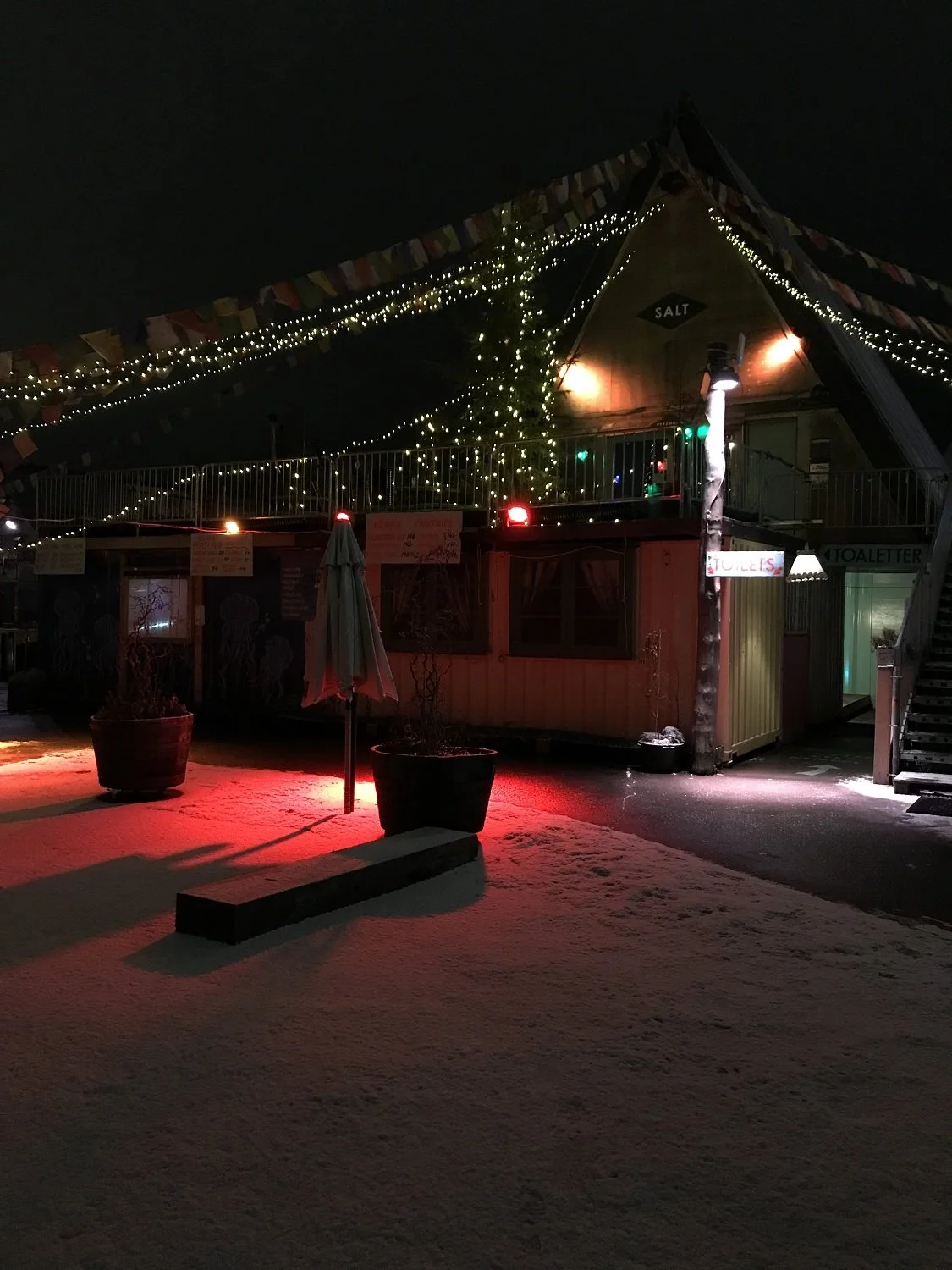 Nighttime scene of a decorated building with string lights and a Christmas tree, snow on the ground, and a red light illuminating the snow.