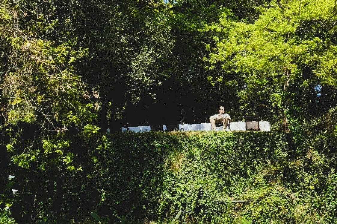 A man sitting on a white outdoor furniture set on a lush green elevated lawn amid trees.