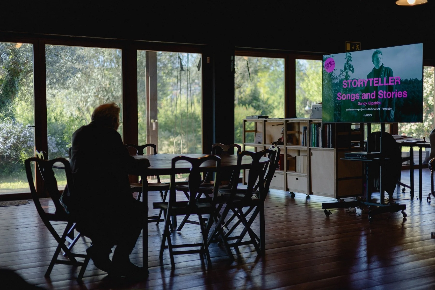 Sandy Kilpatrick sitting at a table in a room with large windows, looking at a screen that displays a presentation titled 'Storyteller Songs and Stories' by Sandy Kilpatrick, with natural outdoor scenery visible through the windows.