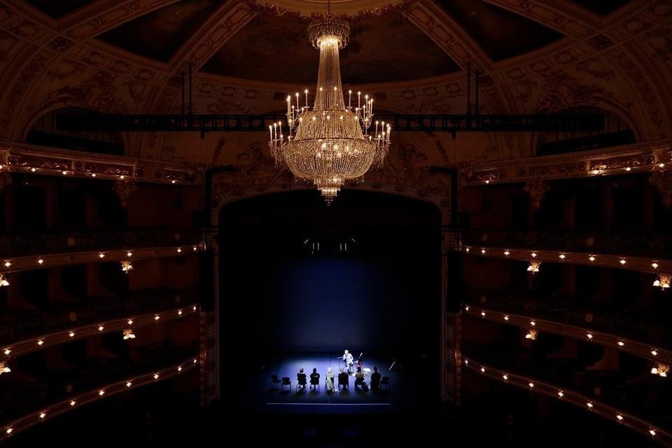 Interior of a theater with a grand chandelier, empty balconies, and a stage with a single performer illuminated by spotlight.