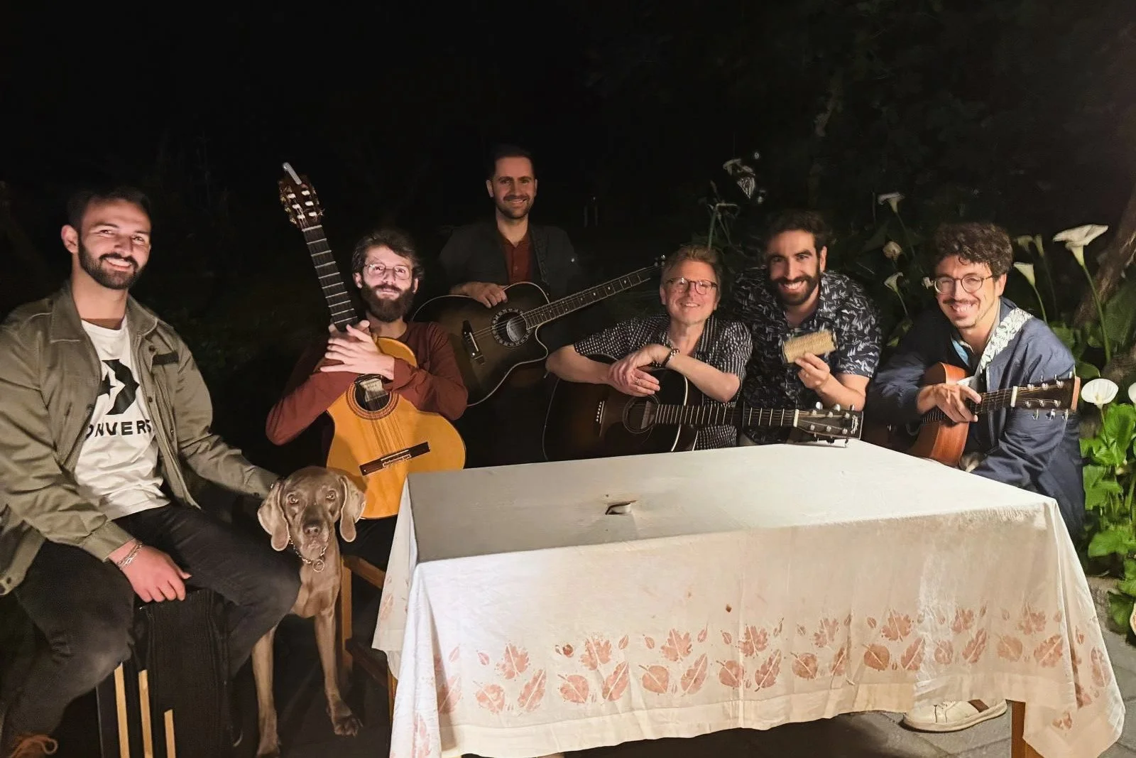 Group of seven men with guitars and a dog gathered around a table outdoors at night, smiling.