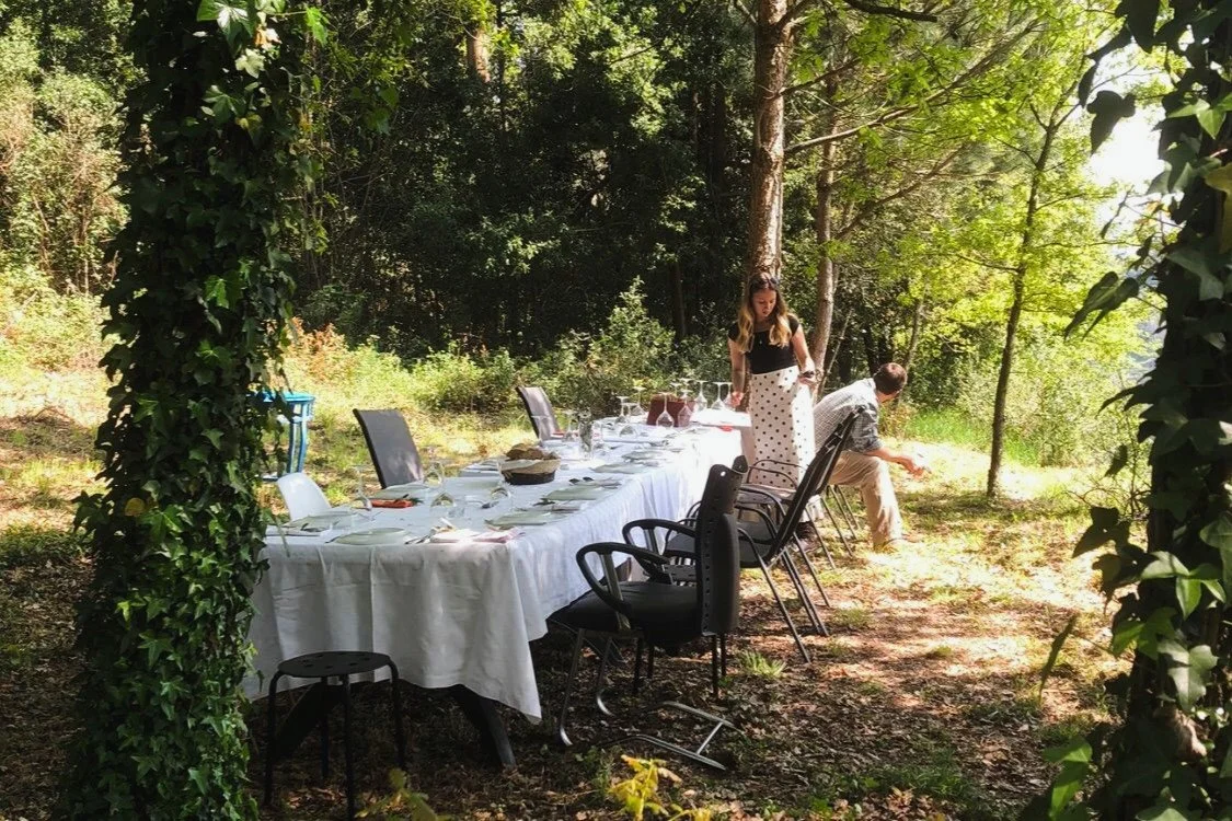 Outdoor dining setup with a long table covered in a white tablecloth, set with glasses, plates, and utensils, surrounded by foliage and trees, with two people preparing or cleaning the area.