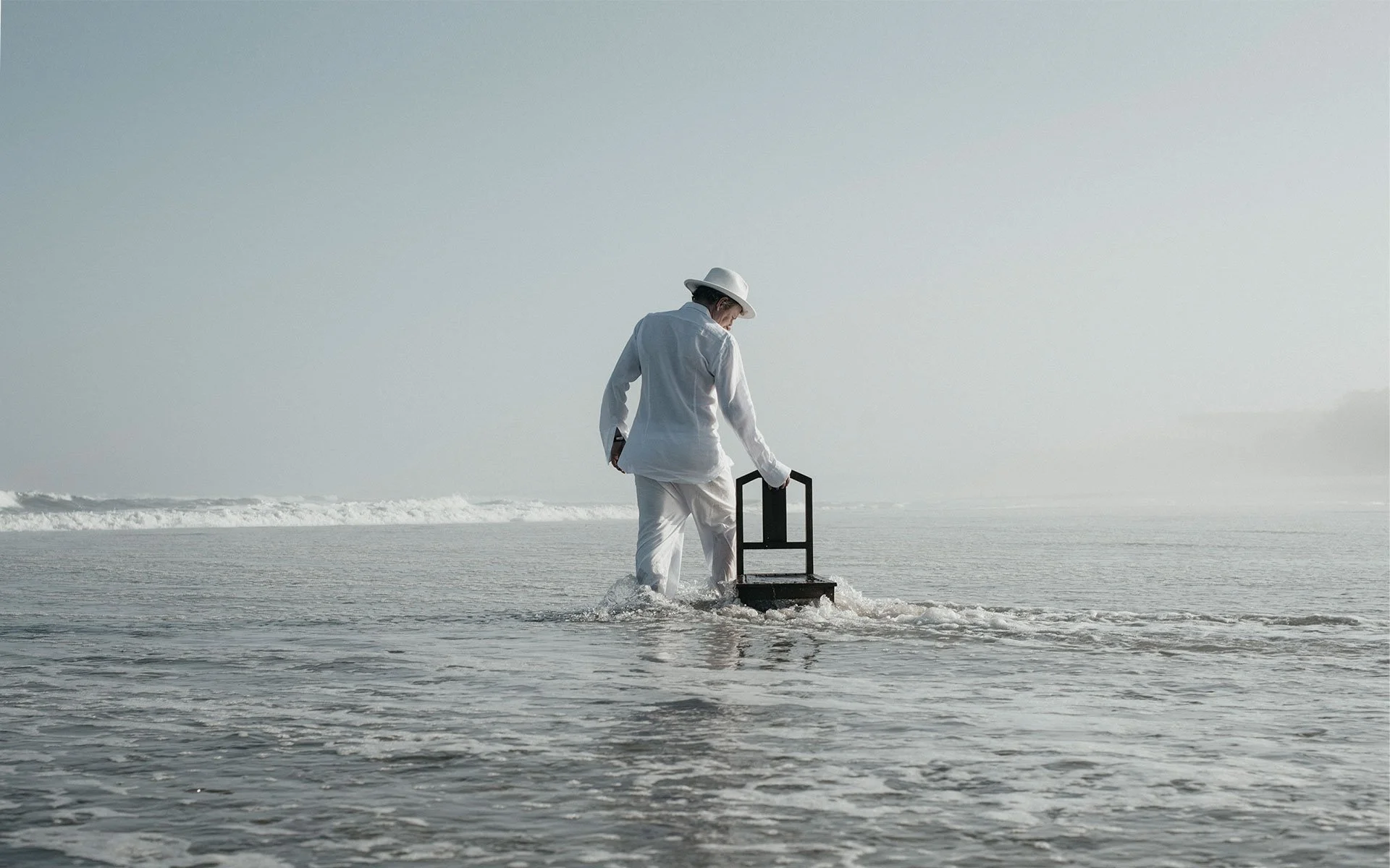 Sandy Kilpatrick dressed in white clothes and a hat is wading in shallow ocean water while holding a black chair. The scene is minimalistic with a vast, calm seascape and cloudy sky.