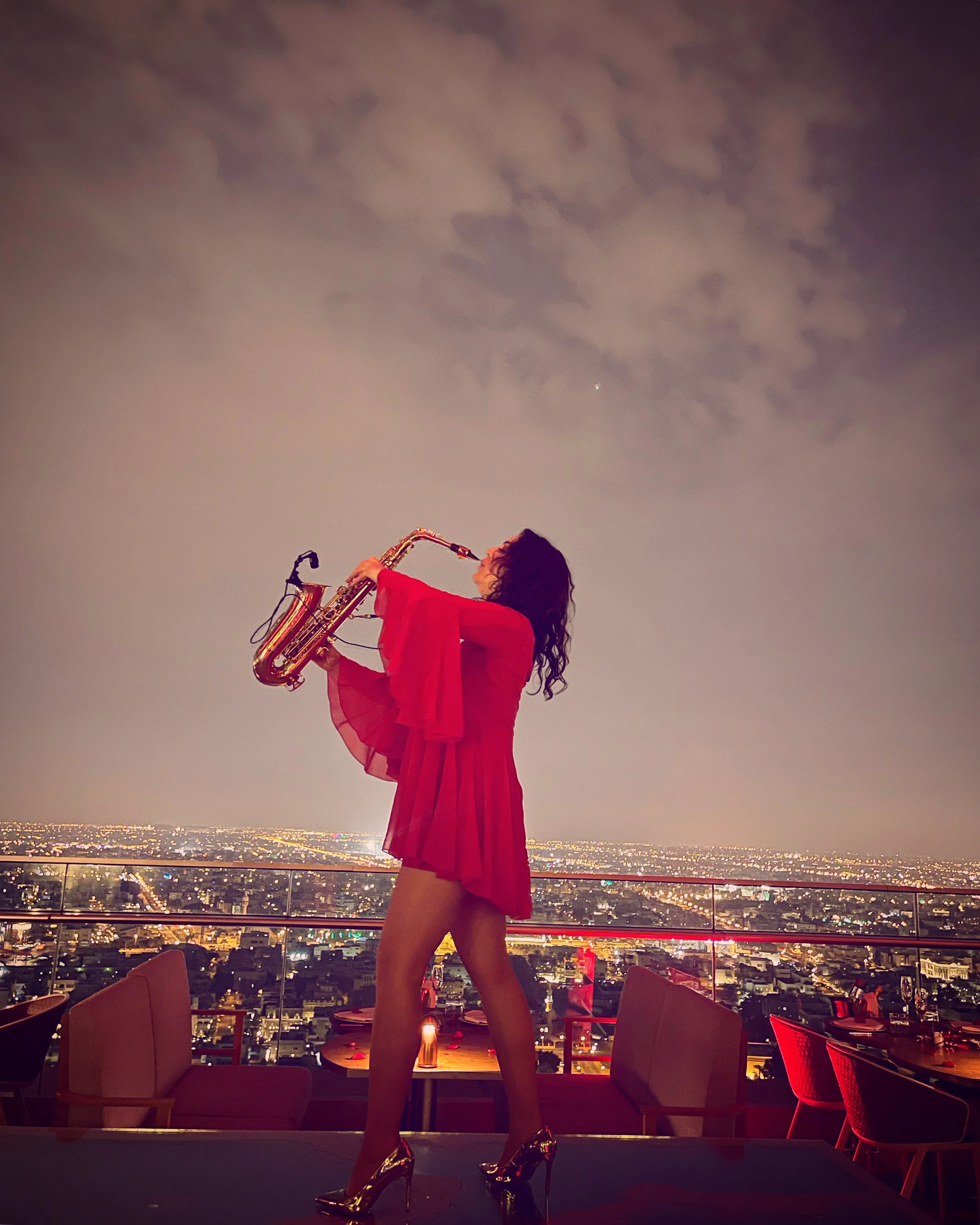 A woman in a red dress and gold high heels playing the saxophone on a rooftop at night, with a city skyline and filled sky in the background.