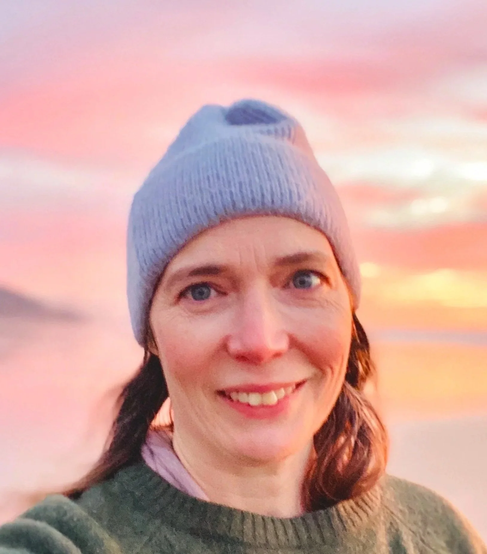 A woman with short curly hair wearing a gray knit beanie and a gray sweater, smiling against a colorful sunset sky with shades of pink and orange.
