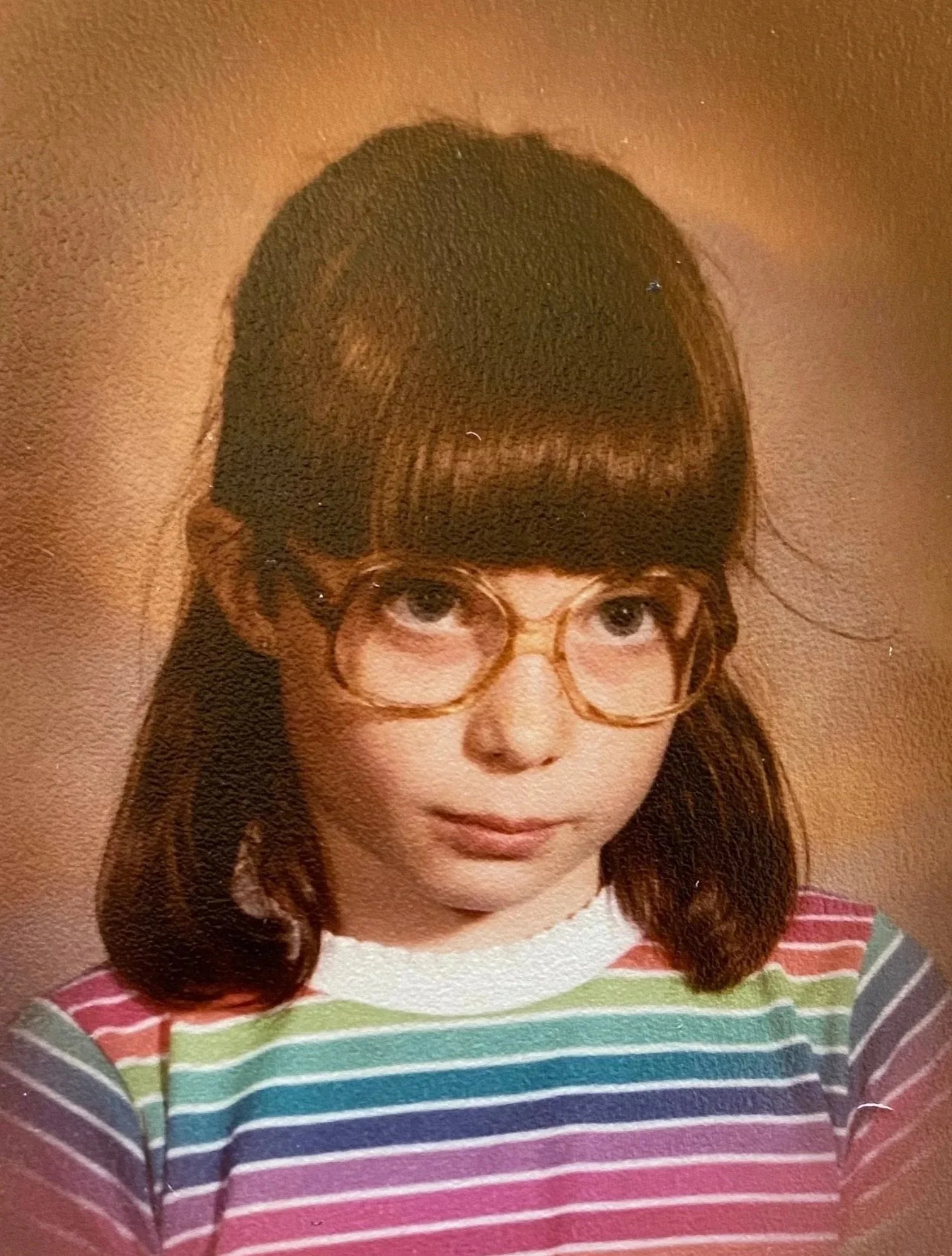 A portrait of a young woman with dark brown hair styled in soft waves, wearing round glasses and a colorful striped top with horizontal rainbow-colored stripes.