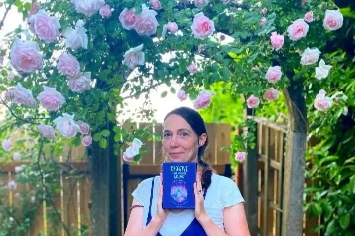 A woman standing outdoors beneath a flowering rose arch, holding a book with a purple cover.