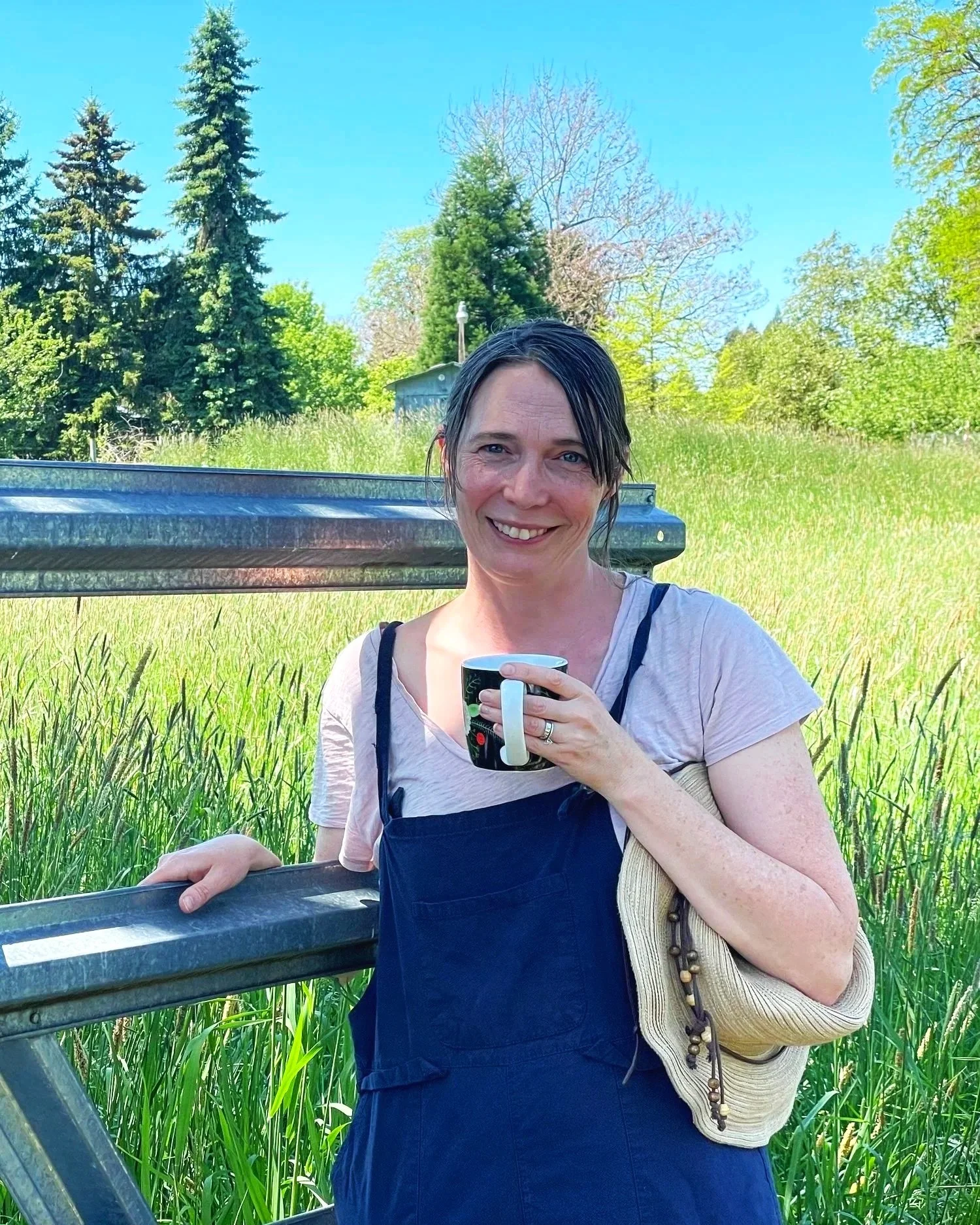 A woman with wet dark hair smiling outdoors on a sunny day, holding a mug, standing near a metal railing in a grassy area with trees and a blue sky in the background.