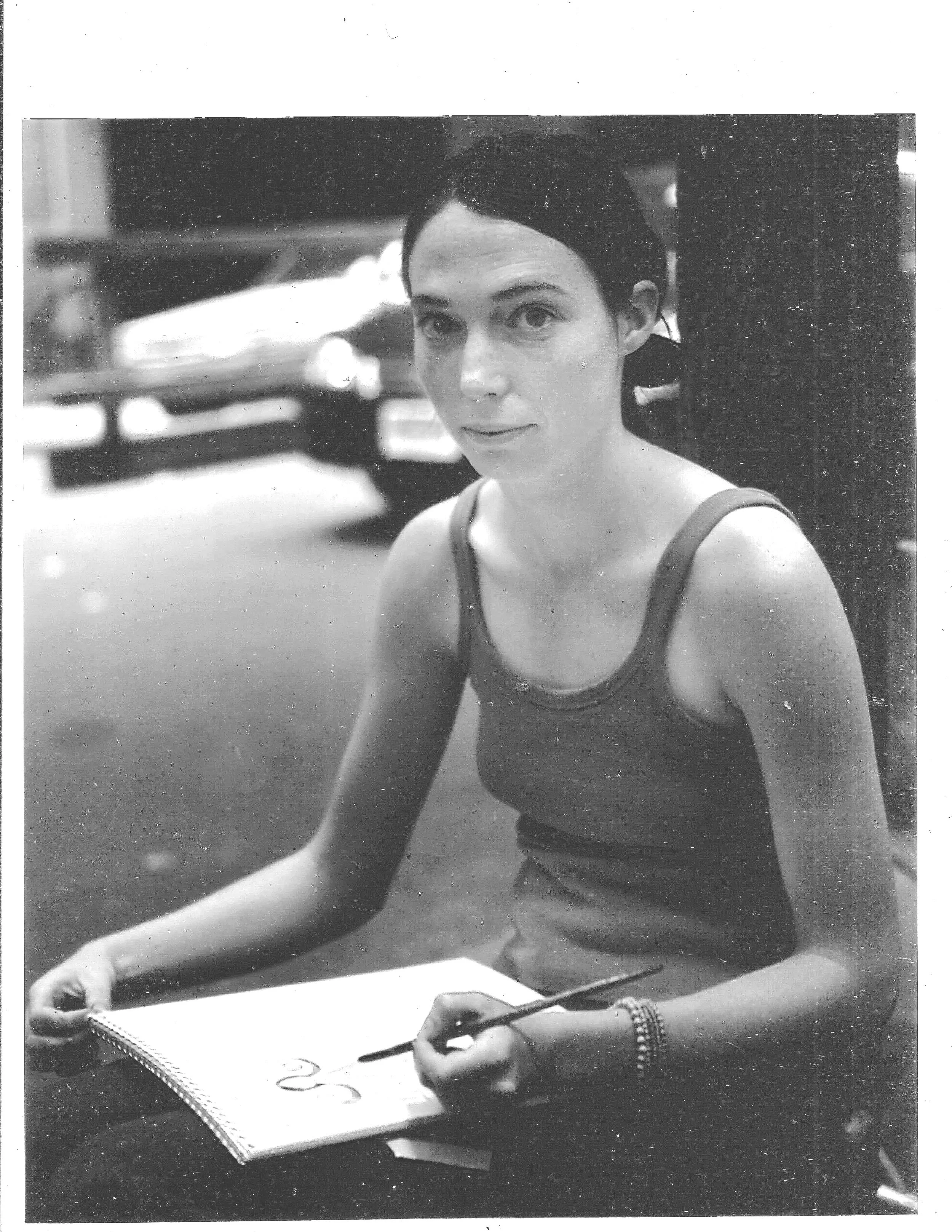 A young woman with dark hair sitting at a desk, writing in a notebook with a pen, wearing a sleeveless top and beaded bracelet, in a black and white photo.