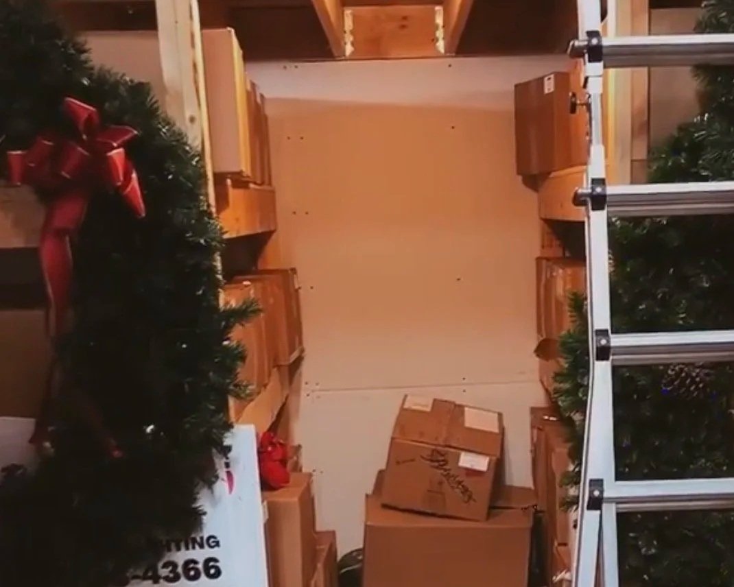 View of a storage room with shelves filled with boxes, holiday wreaths, and a step ladder, with a cardboard box on the floor.