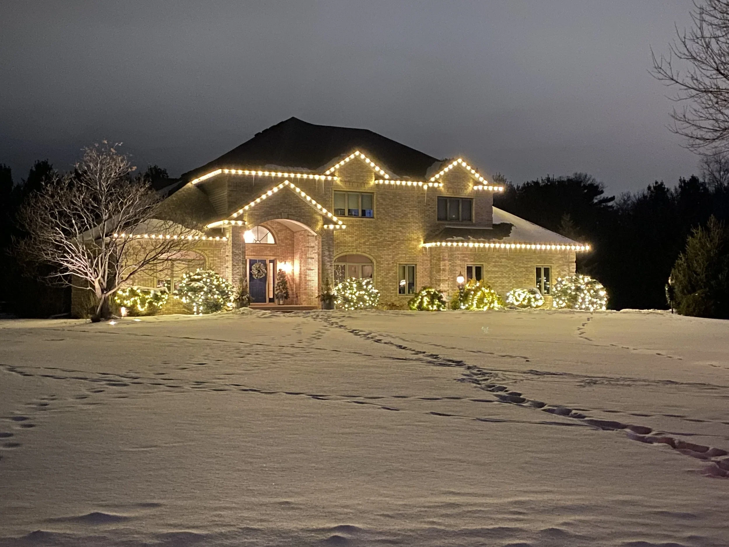 A large brick house decorated with Christmas lights at night, with snow on the ground and footprints leading to the front door.