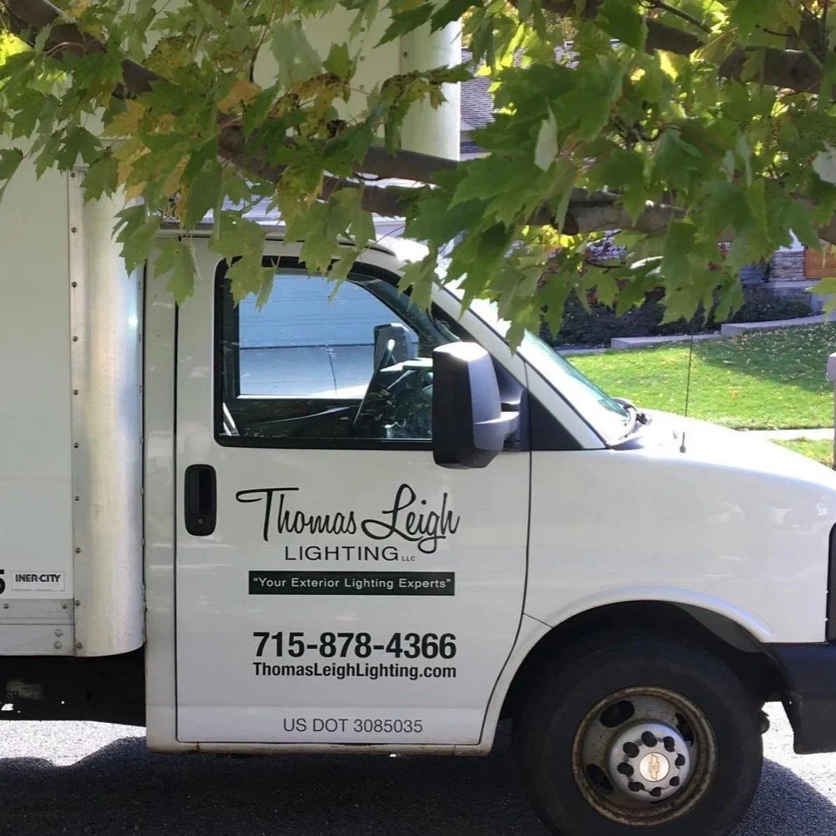 A white utility truck for Thomas Leigh Lighting LLC, with contact information and tagline 'Your Exterior Lighting Experts,' parked on a street, partially obscured by green tree leaves in the foreground.