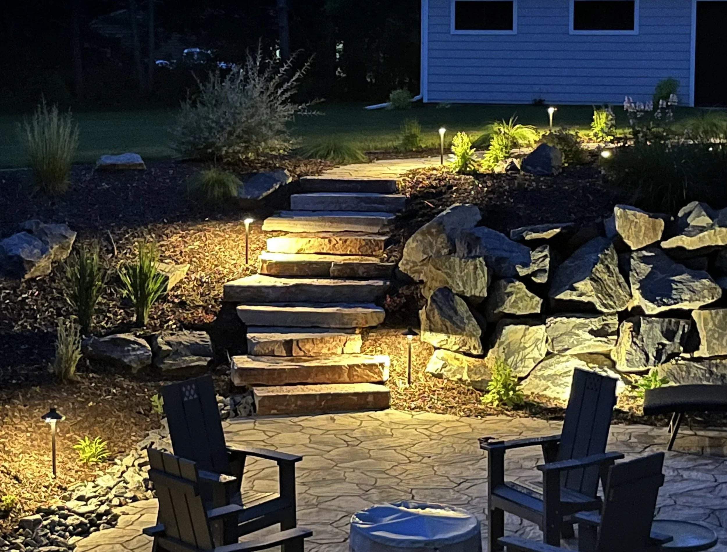 Nighttime backyard scene with stone steps, surrounding rocks, trees, garden lights, and patio furniture.