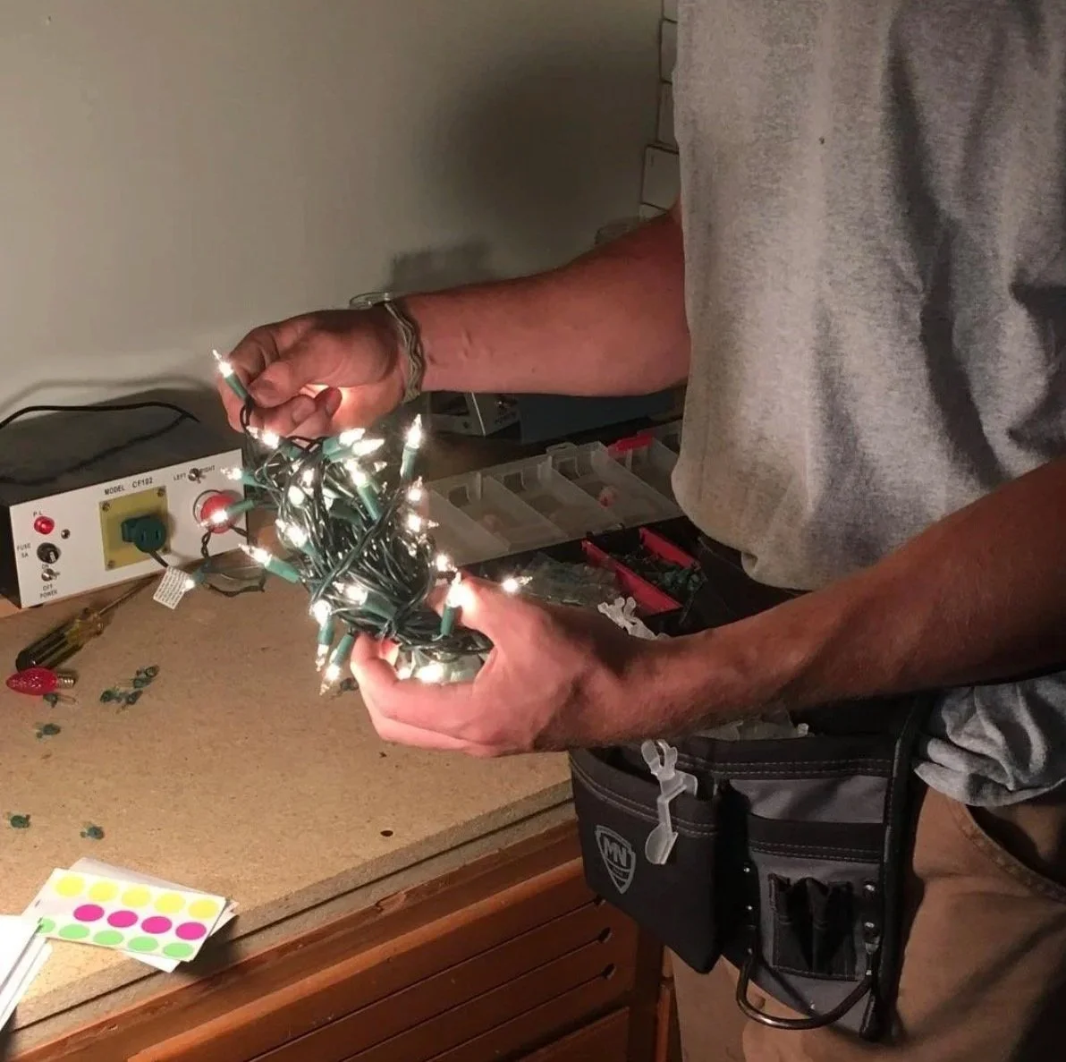 A person holding a tangled bundle of lit Christmas lights over a workbench with tools and wires, wearing a gray T-shirt and a black tool pouch.