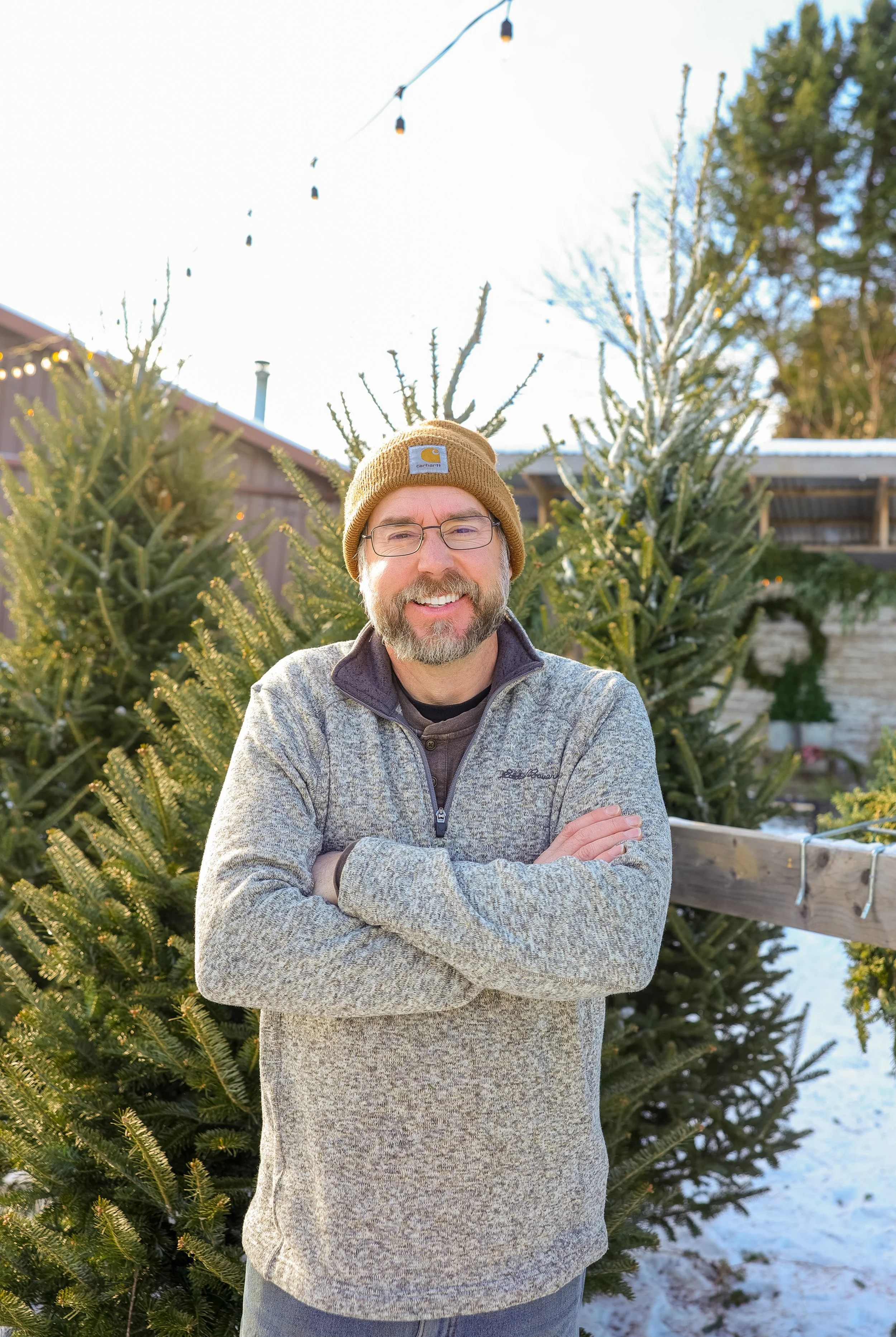 A man smiling with crossed arms standing outdoors in front of Christmas trees on a snowy day, wearing glasses, a brown knit beanie, and a gray fleece jacket.