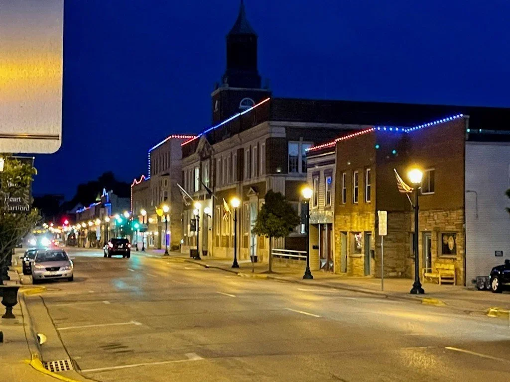Nighttime view of a small-town street decorated with colorful string lights along the building facades, with a few parked cars and street lamps illuminating the sidewalk, and a church tower visible in the background.