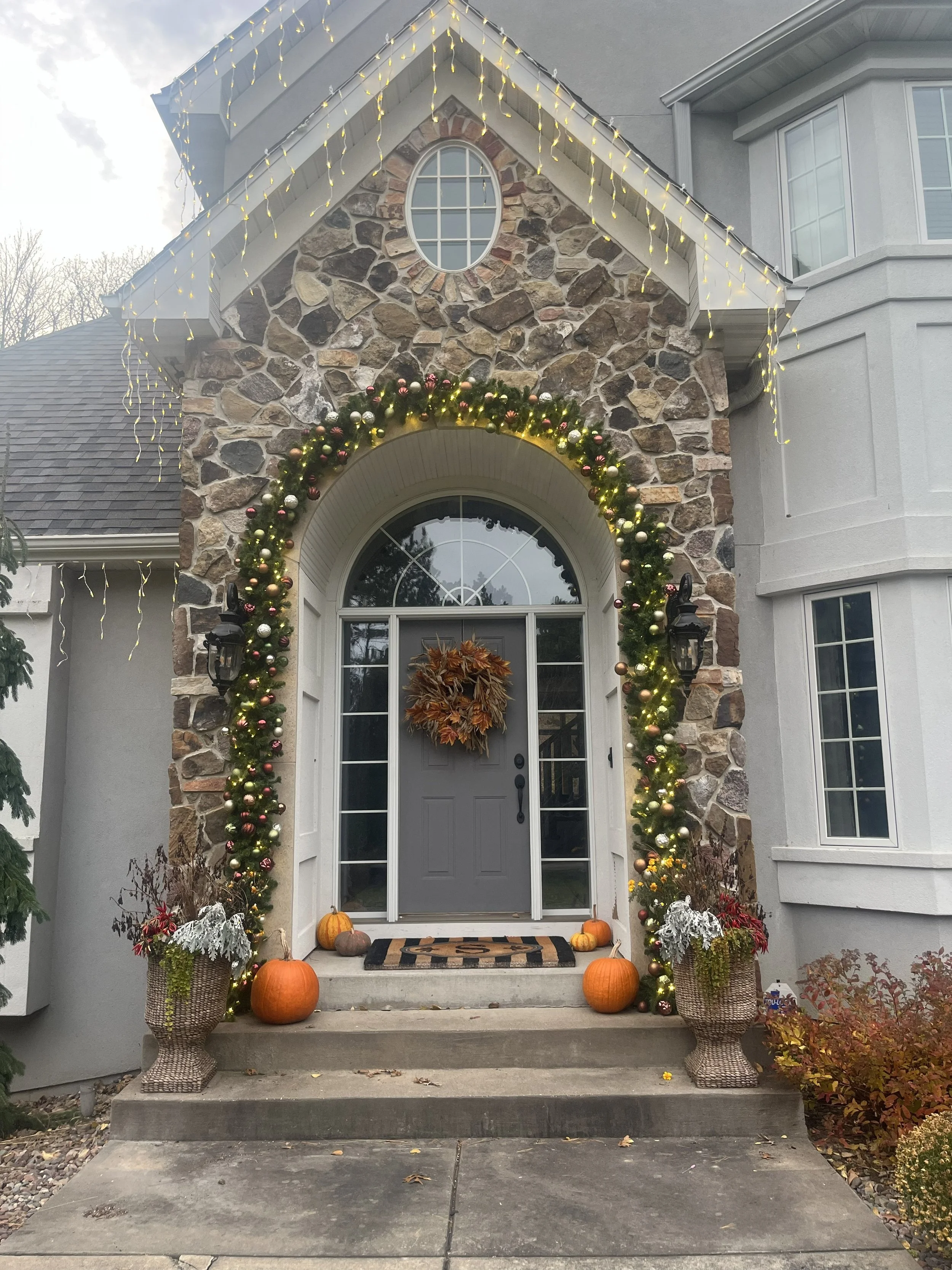 Front entrance of a house decorated for fall with pumpkins, autumn leaves, and floral arrangements on either side of the stairs. A leaf wreath hangs on the gray front door, with string lights and a garland of Christmas ornaments and greenery framing the doorway.