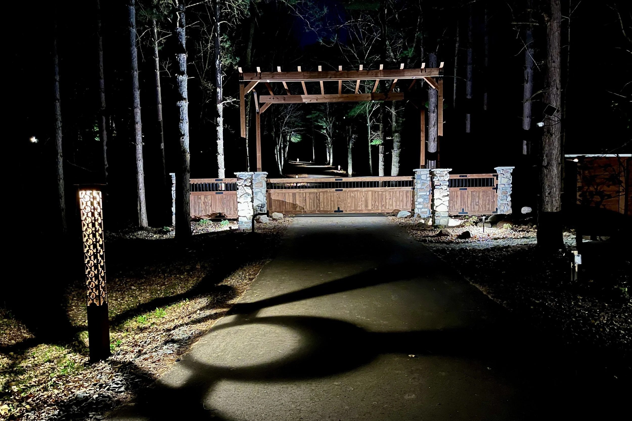 Night view of a wooden Gate and path leading through a forest with tall trees, illuminated by outdoor lights.