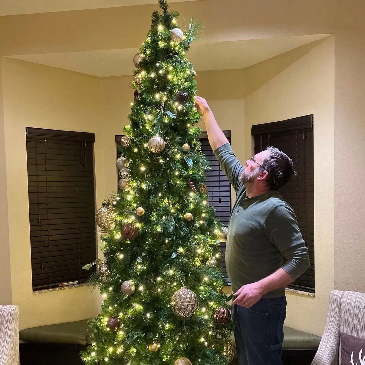 A man decorating a tall Christmas tree with white and gold ornaments and lights in a room with yellow walls and black window blinds.