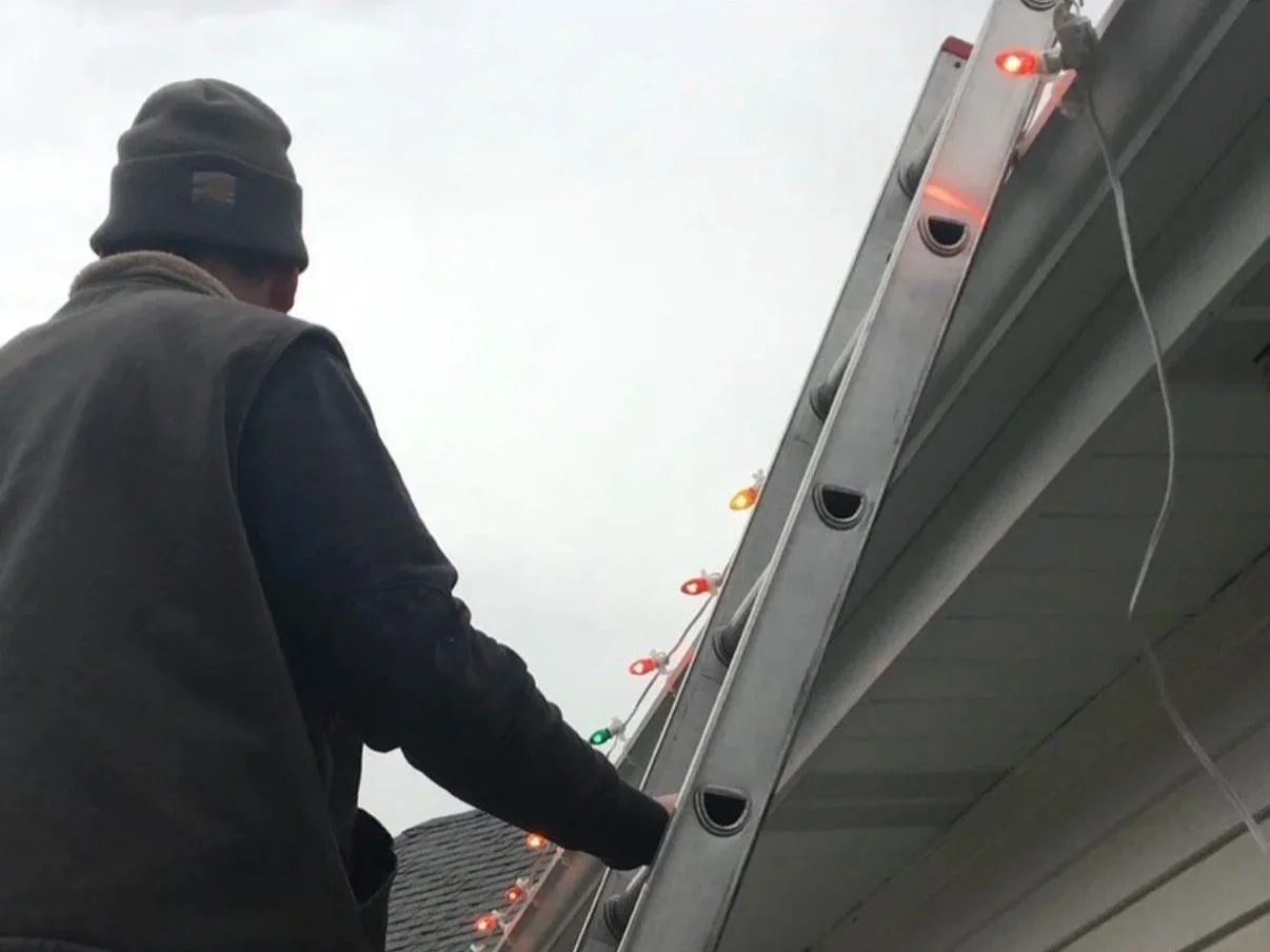 A man adjusting or installing Christmas lights on the gutter of a house roof using a ladder, with the sky in the background.