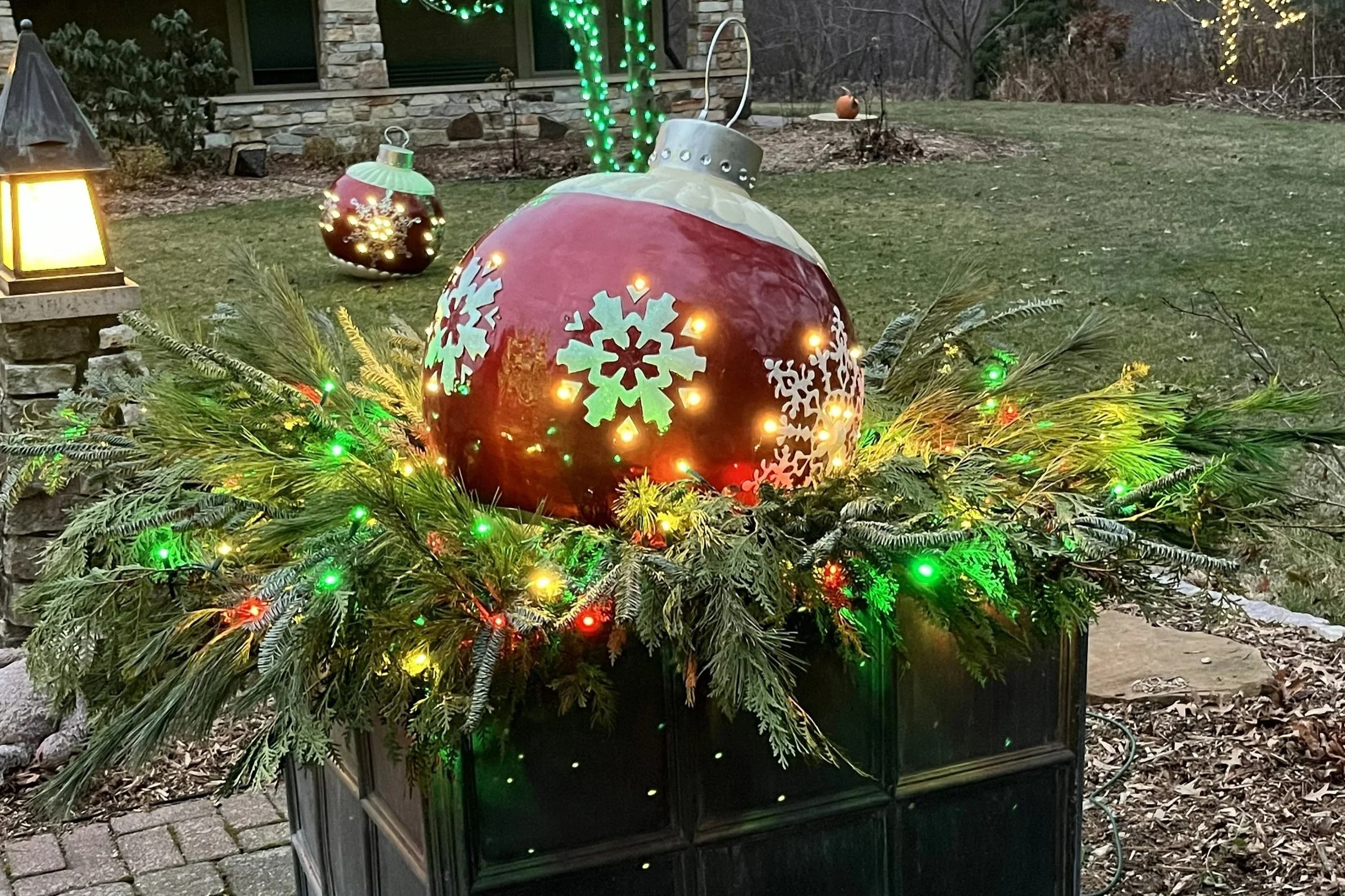 Large red Christmas ornament decorated with white snowflakes, surrounded by green pine branches with multicolored string lights, placed on a black box outdoors during evening.