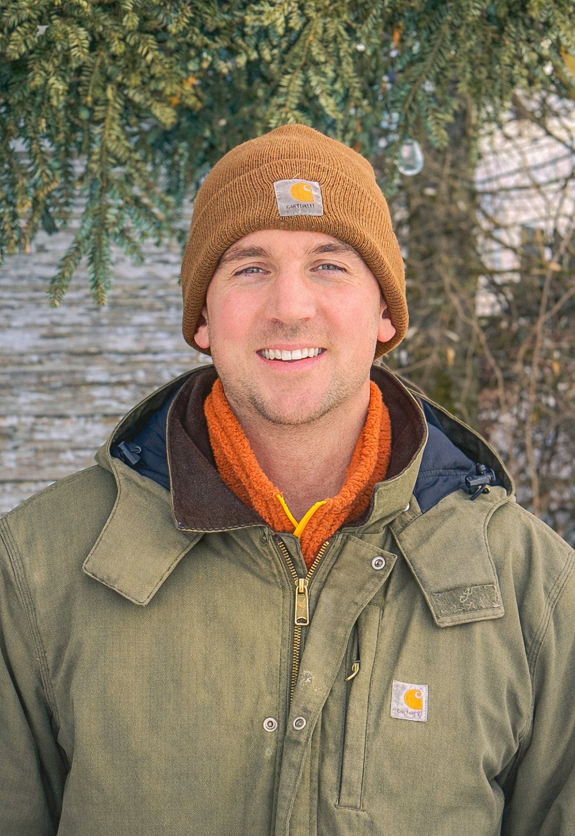 Sawyer smiling outdoors wearing a brown Carhartt beanie, orange fleece jacket, and green jacket in front of a snowy background with trees.