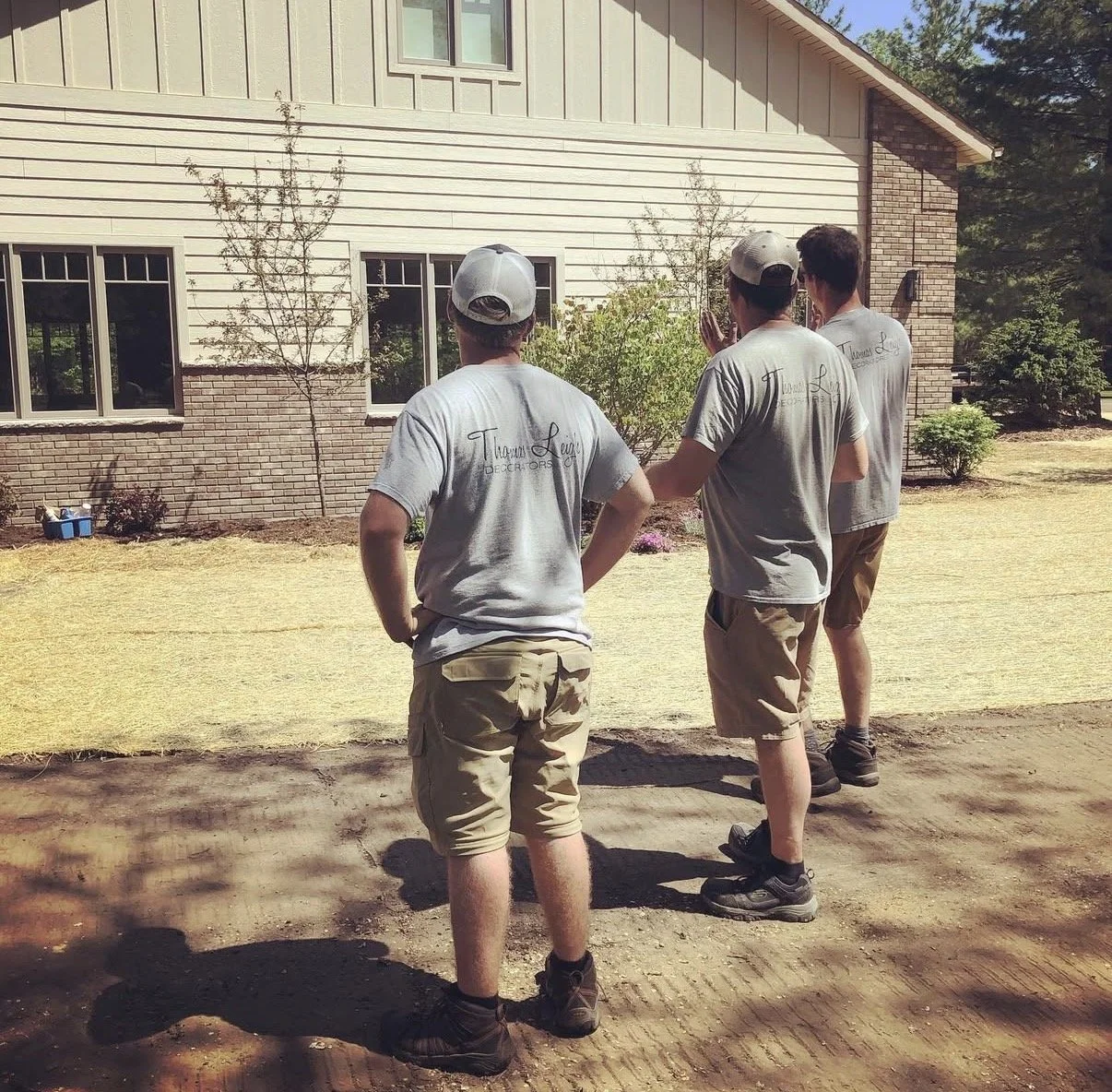 Three men in matching gray T-shirts and shorts are working outside a house, possibly laying turf or involved in landscaping.