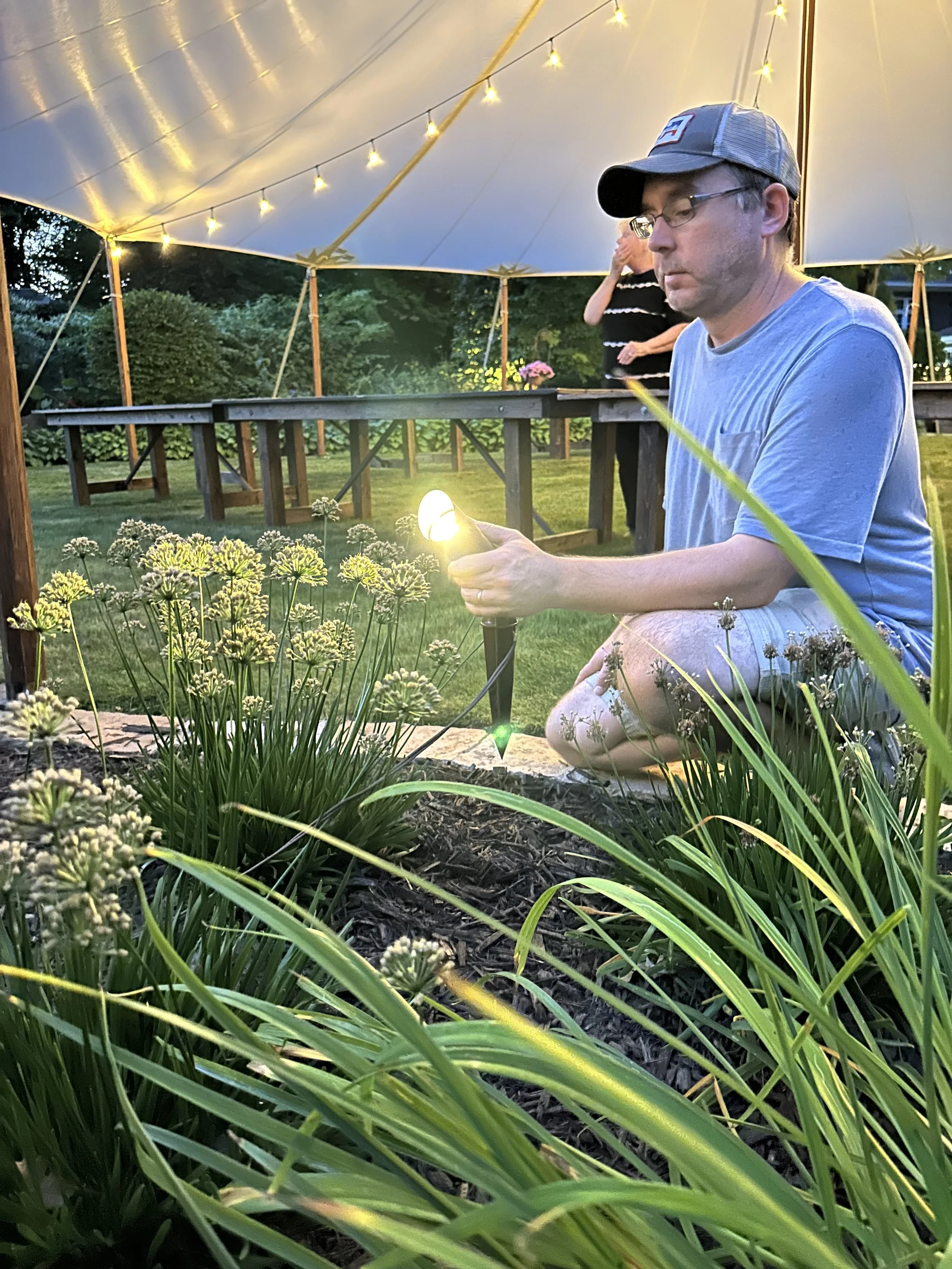 A man kneeling beside flowers in a garden at dusk, holding a small flashlight.