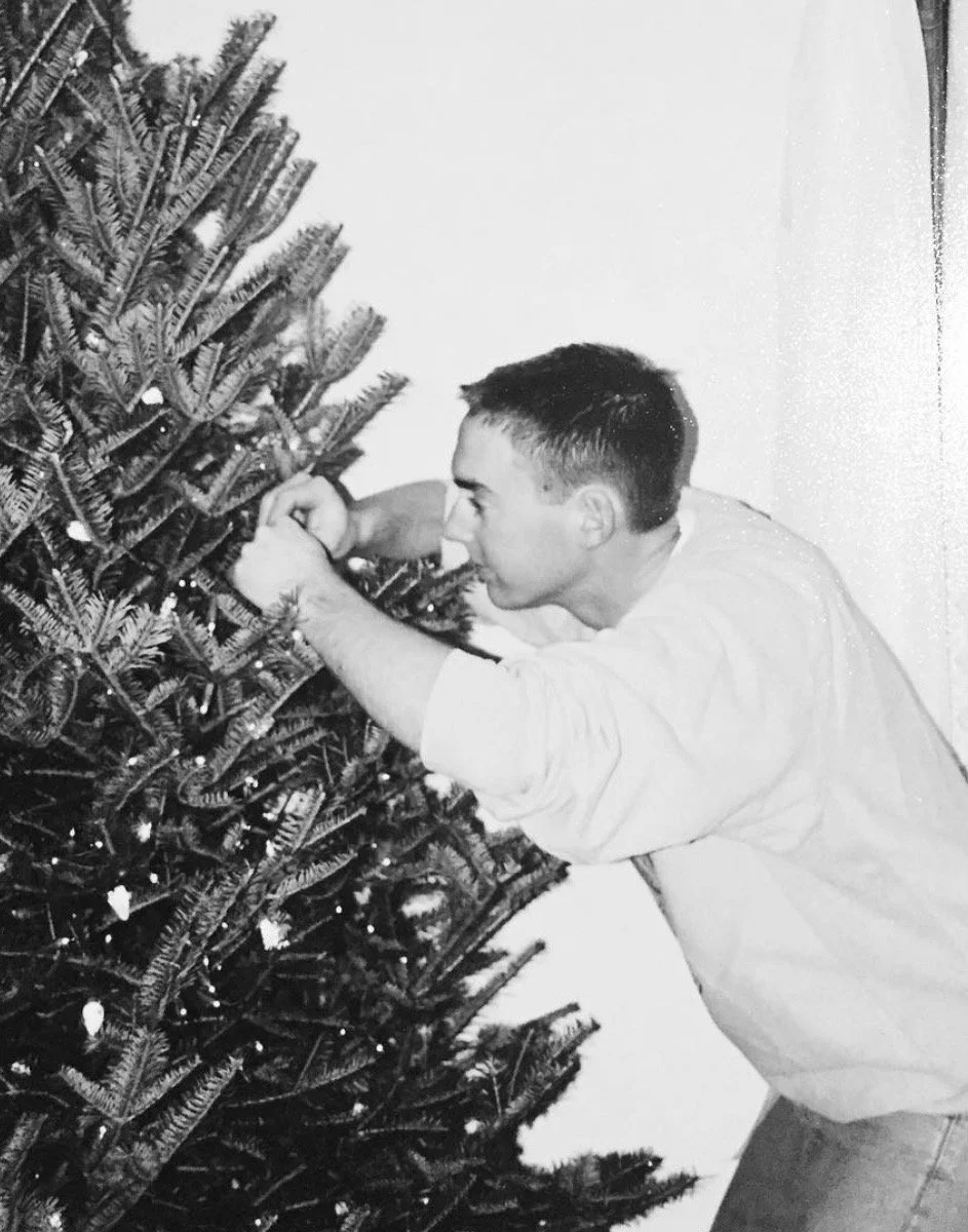 A young man decorating a Christmas tree with ornaments in a black-and-white photo.