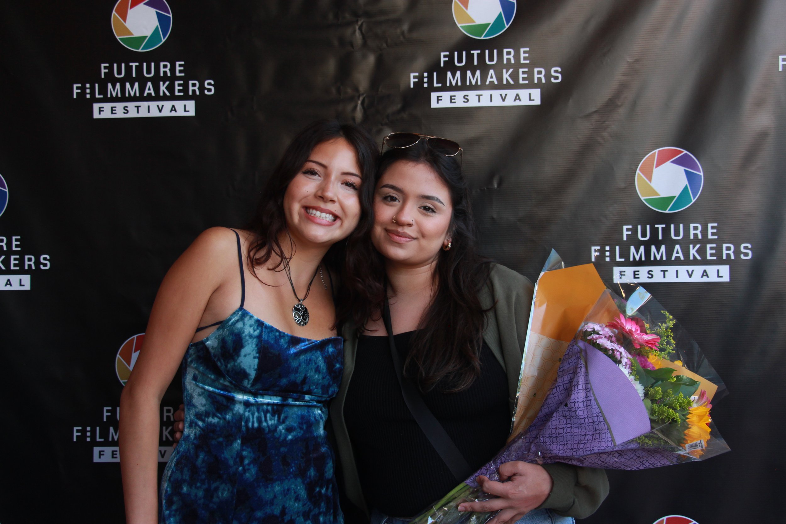 Two women smiling at an event, holding a bouquet of flowers, standing in front of a black backdrop with the 'Future Filmmakers Festival' logo repeated across it.