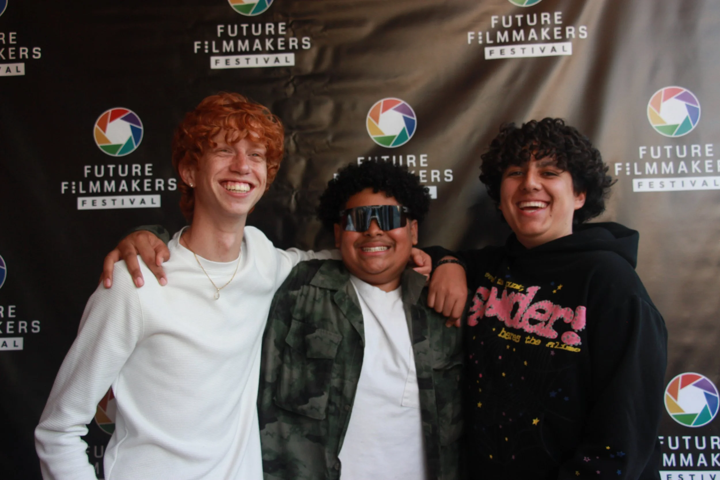 Three young men smiling with their arms around each other in front of a backdrop with the Future Filmmakers Festival logo.