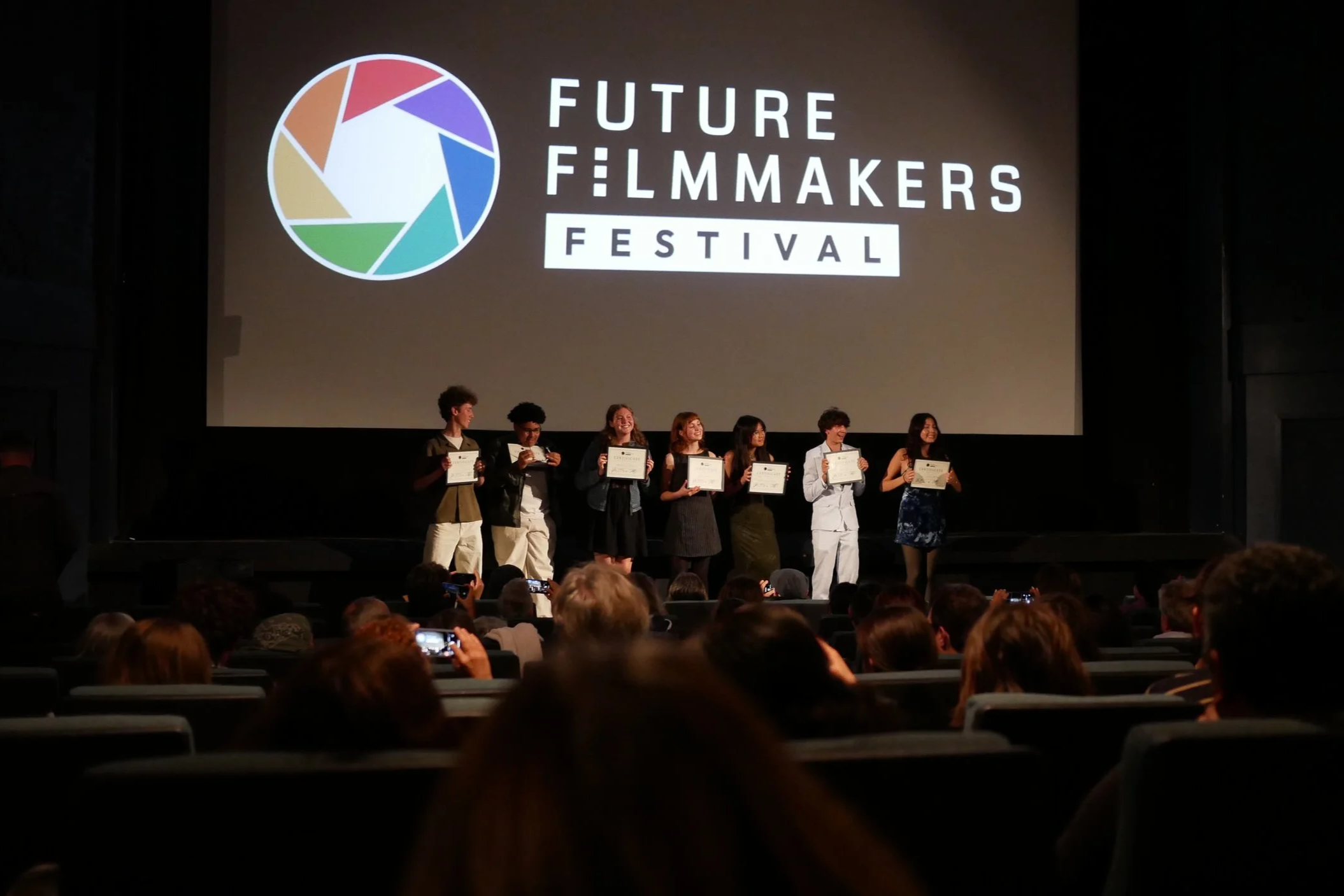 Group of people on stage at Future Filmmakers Festival receiving awards, with audience taking photos, large screen displaying festival logo in background.