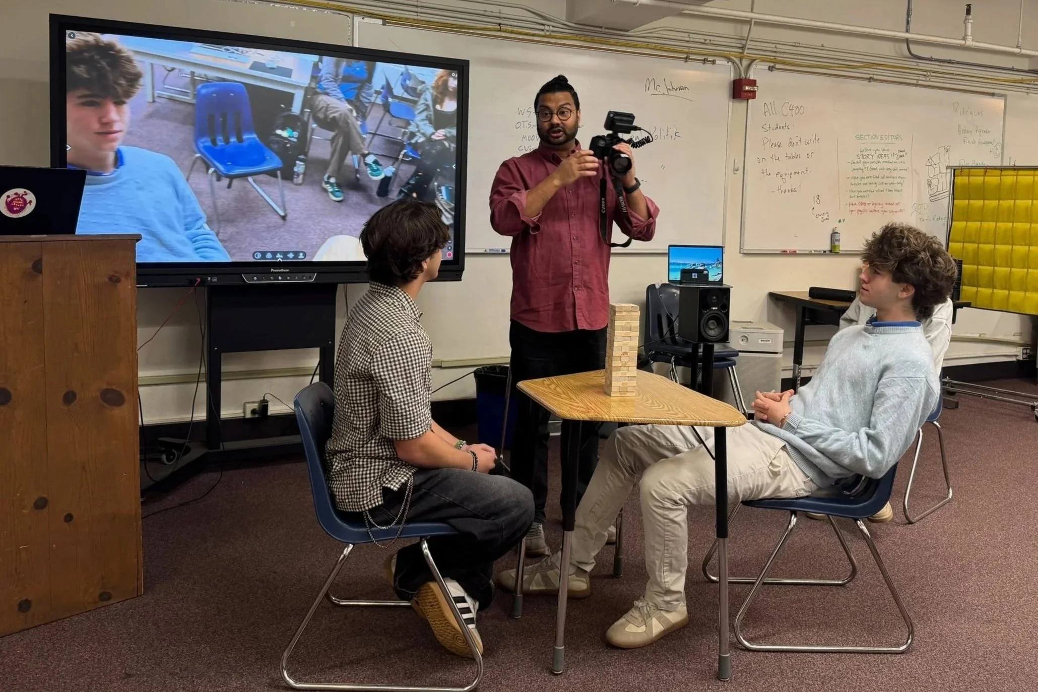 Two students sit at a table in a classroom, playing a game of Jenga, while a teacher or videographer records them with a camera. A large screen behind them shows a video call with more students. The classroom has a whiteboard with writing and drawing