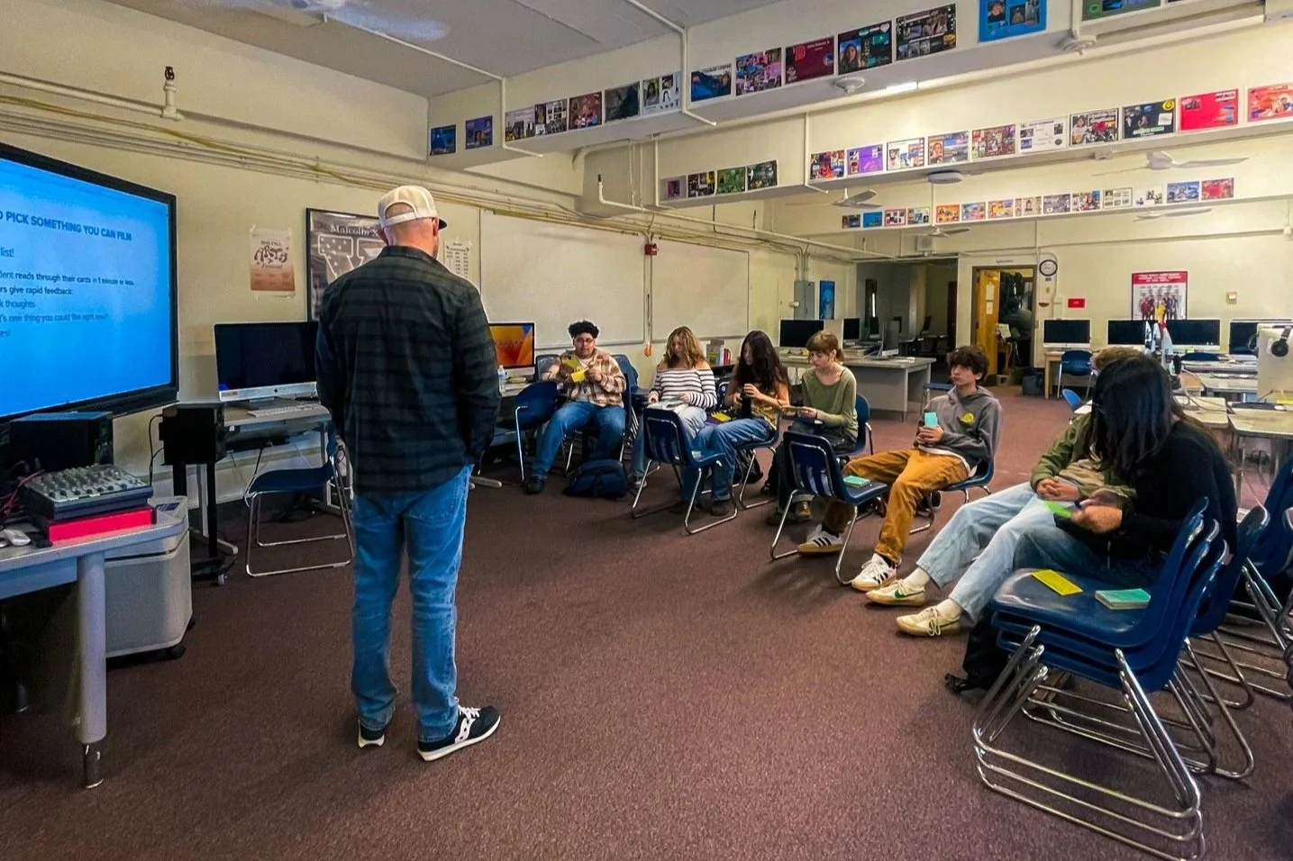 A group of students seated in chairs in a classroom or computer lab, using their phones, with an instructor standing at the front near a large screen displaying instructions.