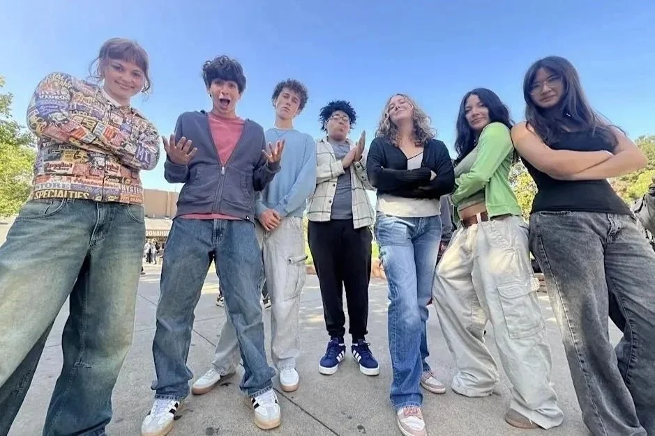 Group of seven teenagers standing outdoors in front of a building, posing playfully for the camera on a clear day.