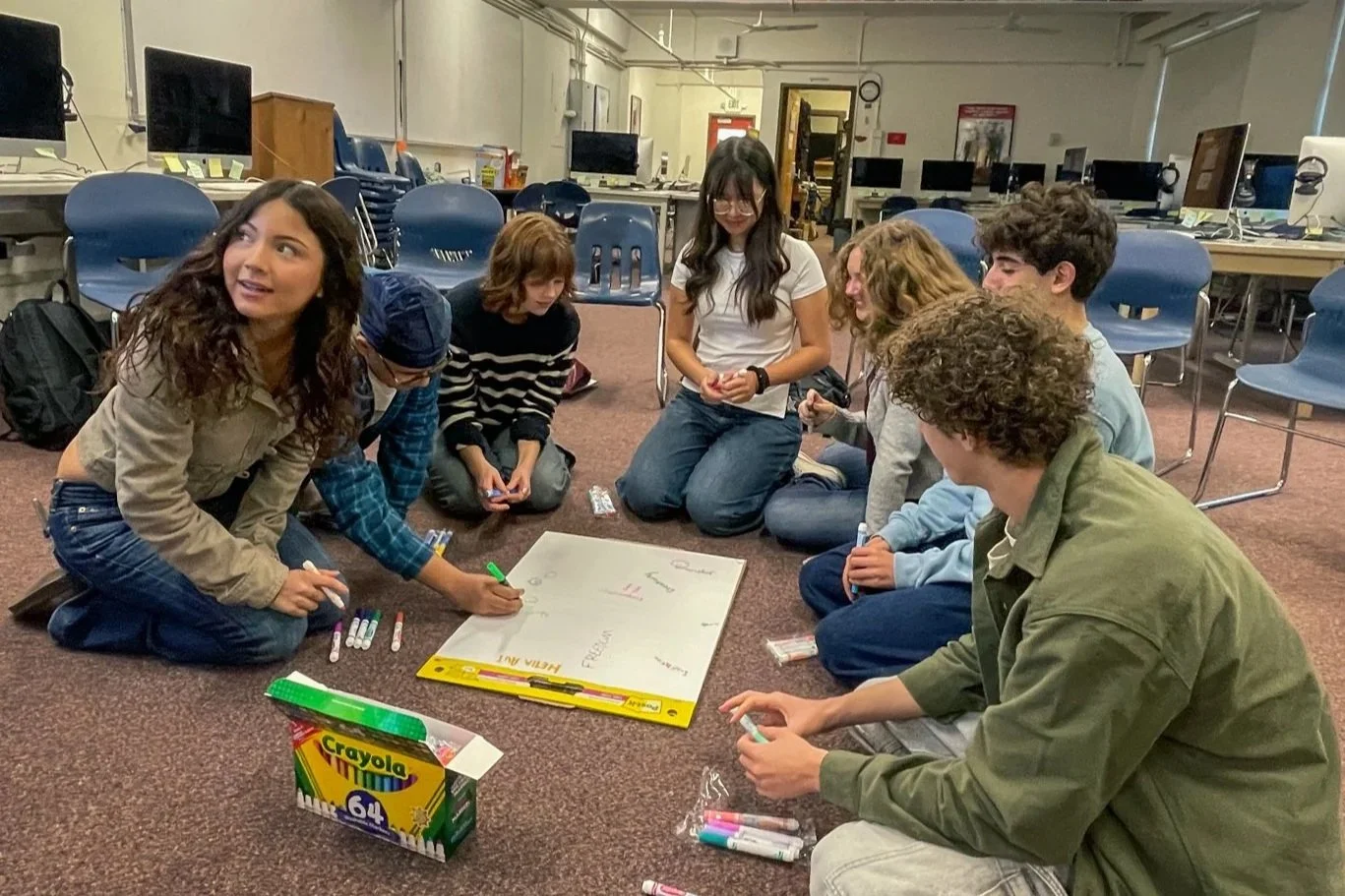 Group of seven teenagers sitting on the floor in a classroom, gathered around a whiteboard with markers, engaging in a collaborative activity.