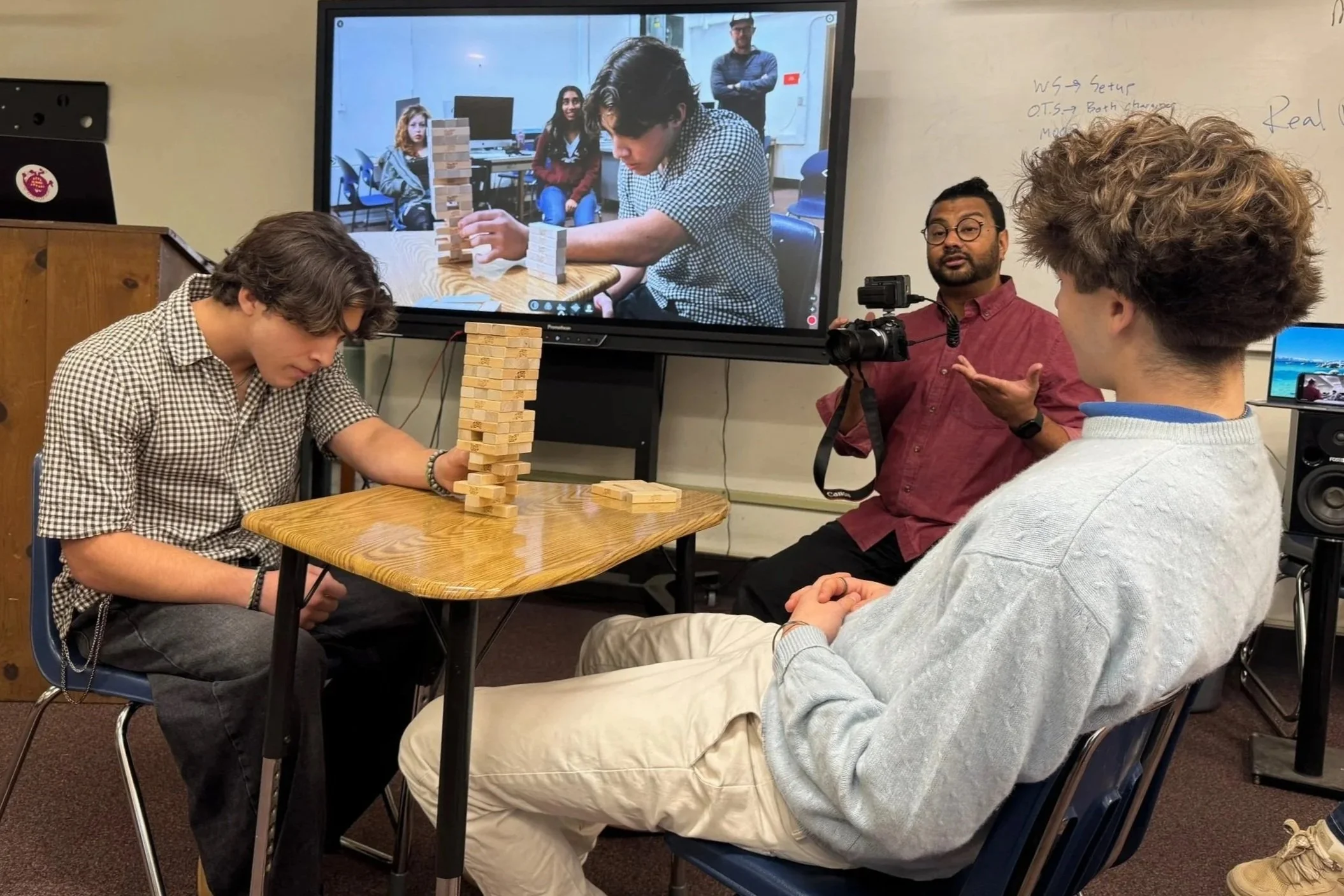 A classroom scene with two students playing Jenga at a table, while a teacher and a student look on. A large screen shows a close-up of the student playing Jenga, with other students in the background.