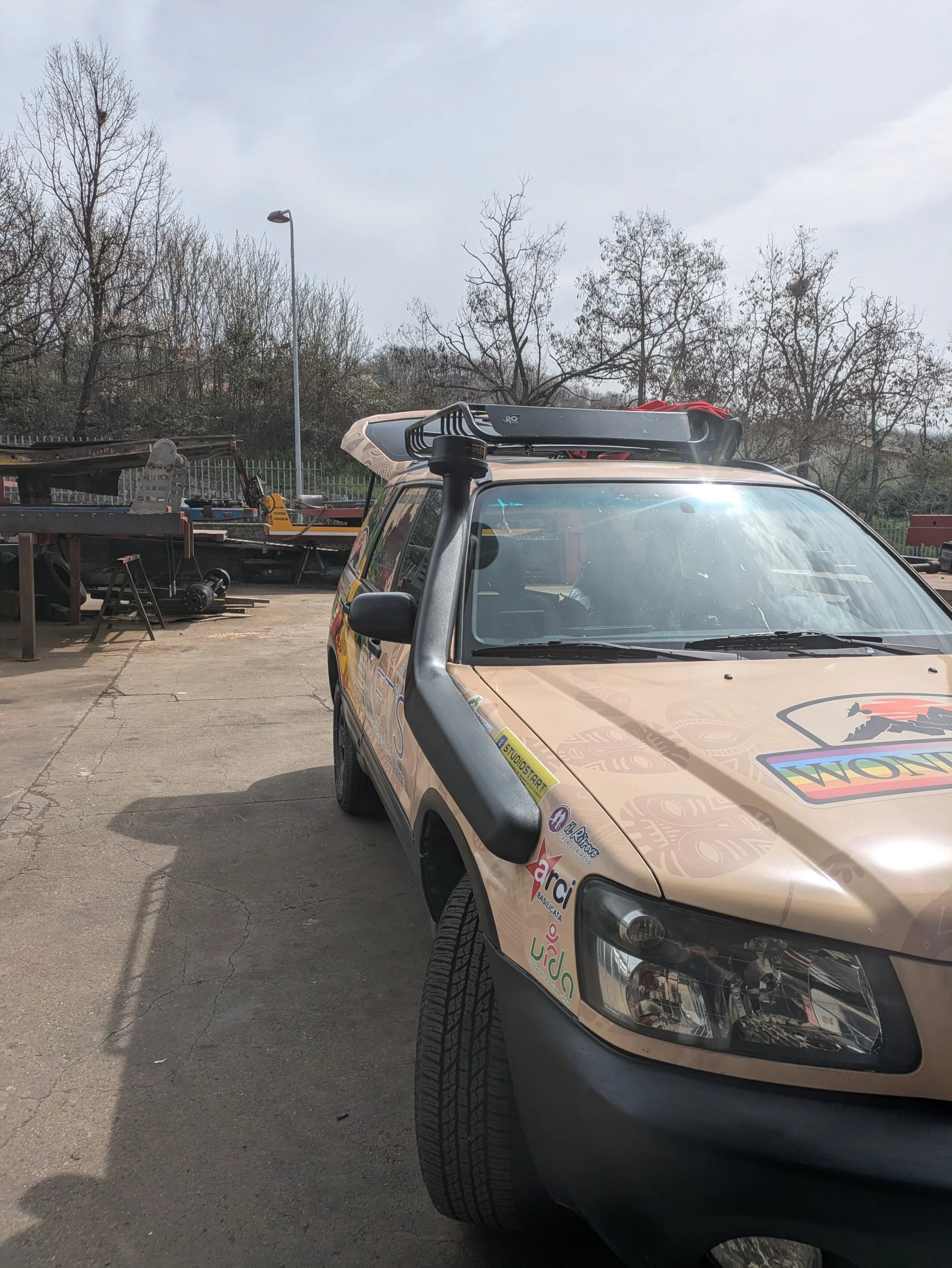 Auto con adesivi pubblicitari parcheggiata in un'area aperta, con barche in costruzione o riparazione sullo sfondo e alberi spogli.