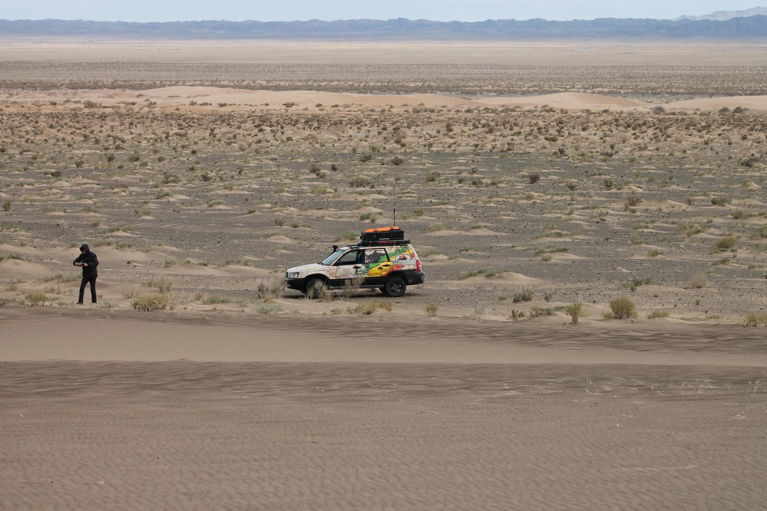 Un'auto da turismo decorata nel deserto con un uomo che la controlla, in un paesaggio di dune e arbusti sparse.