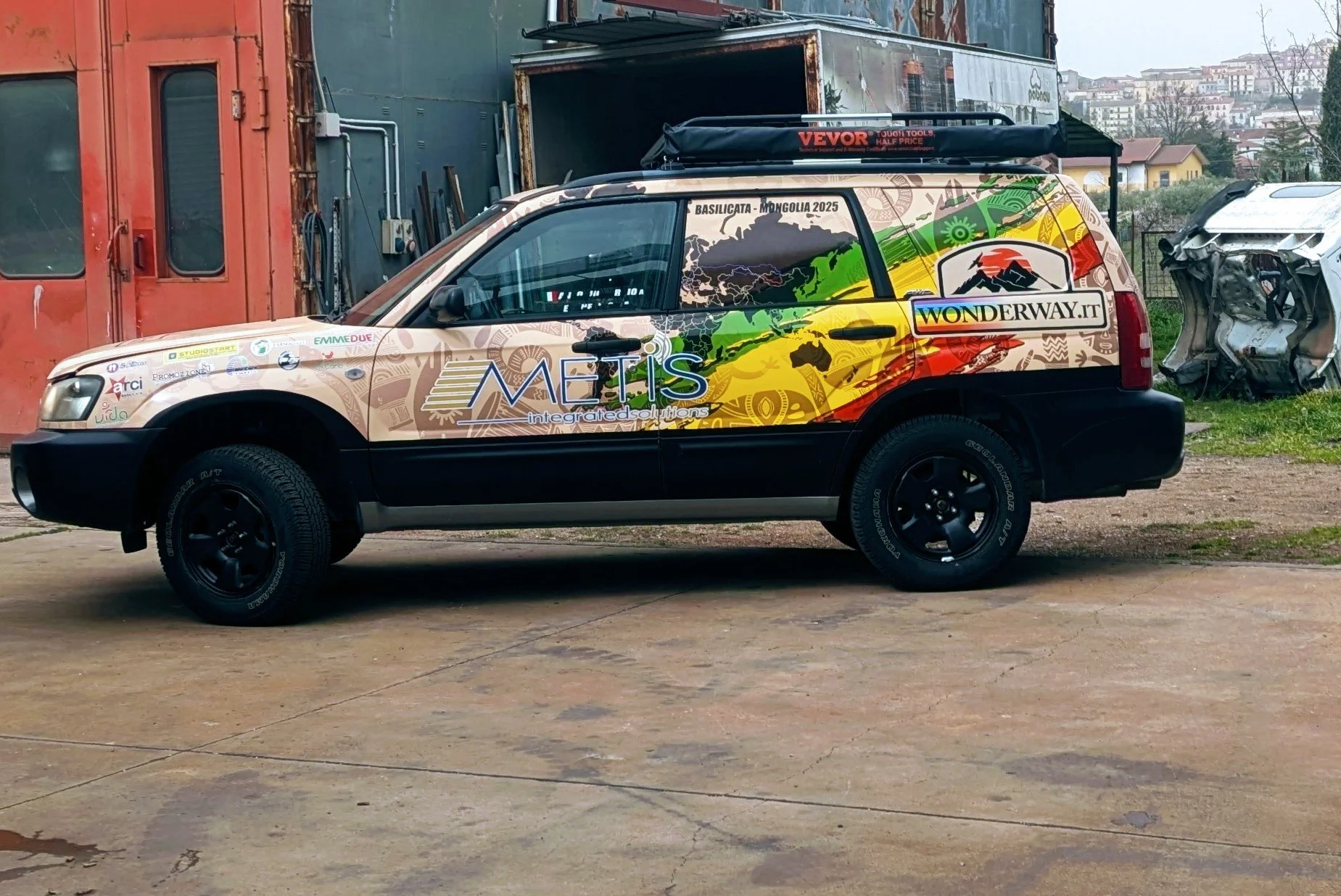 Automobile con decorazioni colorate e logo di METIS, sponsorizzata da WonderWay.it, parcheggiata su una superficie di cemento, con uno sfondo di strutture industriali e edifici residenziali.