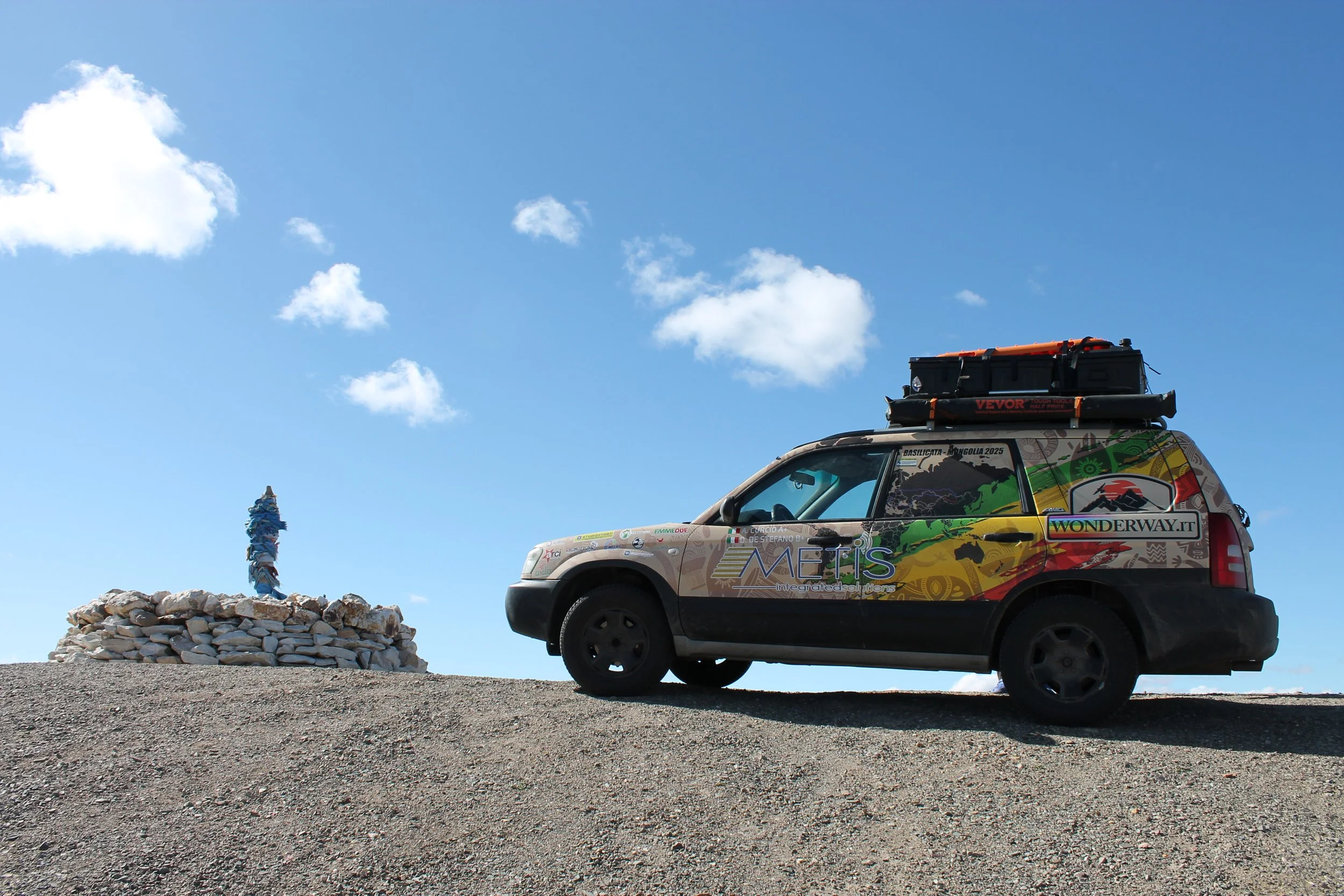 Auto colorata con adesivi e il logo 'WONDERWAY.IT' parcheggiata su un terreno polveroso sotto un cielo azzurro con alcune nuvole, con un cumulo di pietre e un totem decorativo in pietra sulla sinistra.