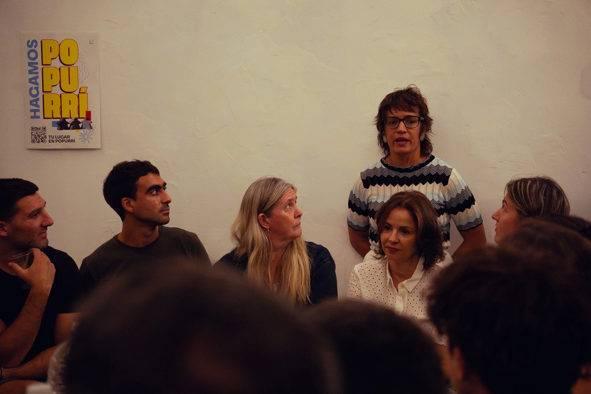 Una mujer de cabello rizado y gafas está hablando delante de un grupo de personas sentadas, algunas mirándola atentamente.