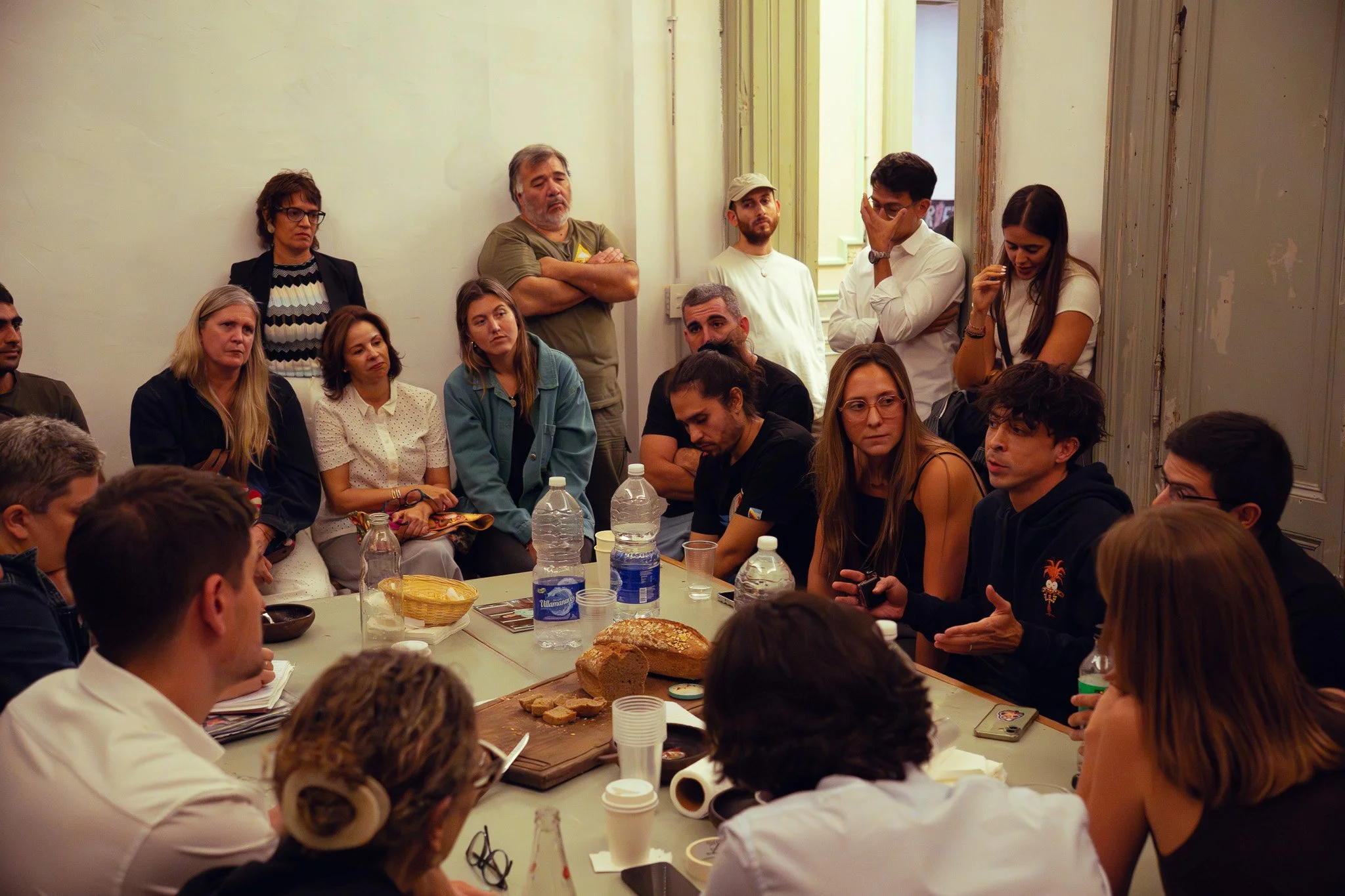 Reunión de personas en una mesa con alimentos y bebidas, discutiendo o participando en una conversación en una sala cerrada.