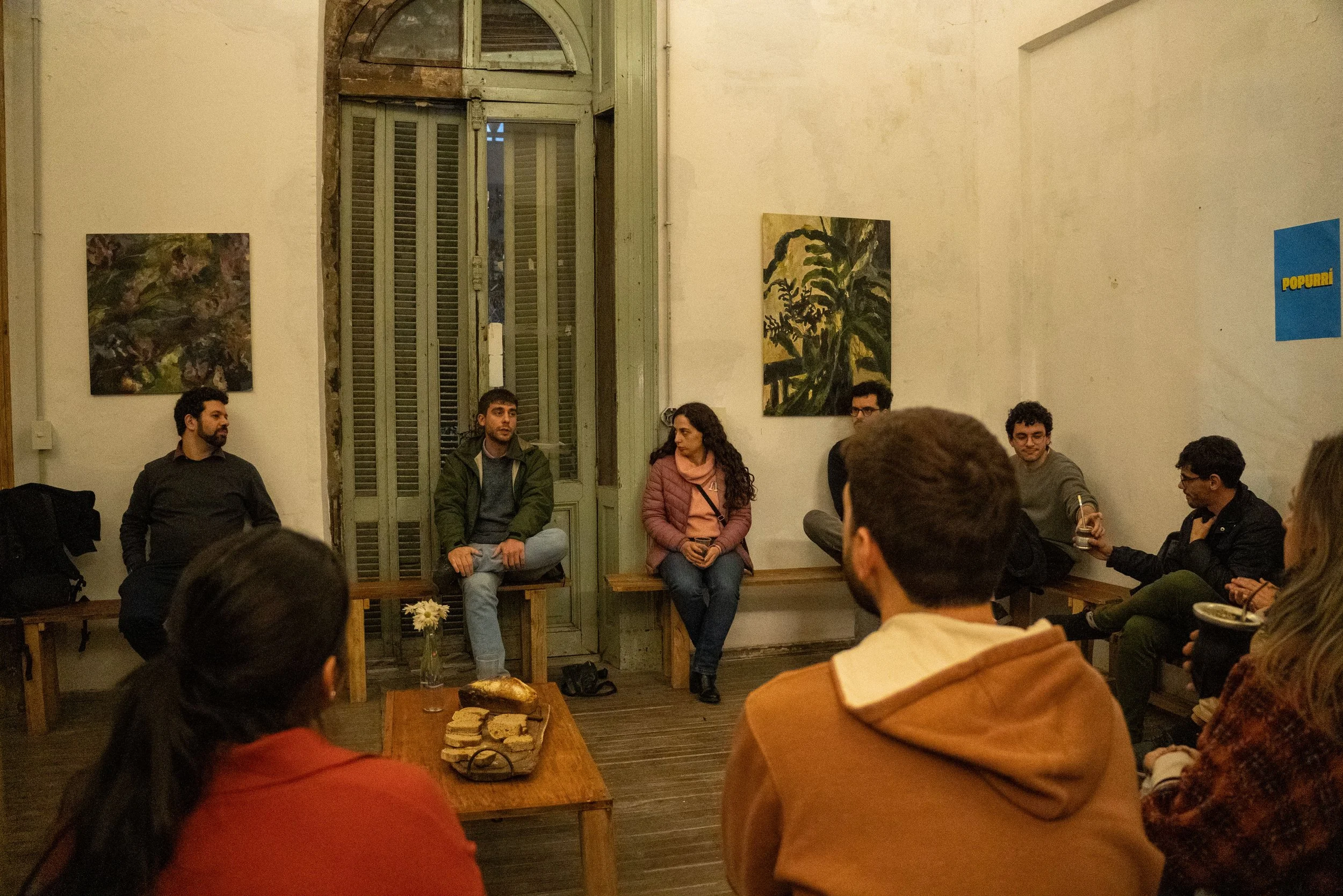 Un grupo de personas sentadas en una sala de reuniones, participando en una charla o discusión. En la pared hay varias pinturas y un cartel azul con la palabra "POPURRI".