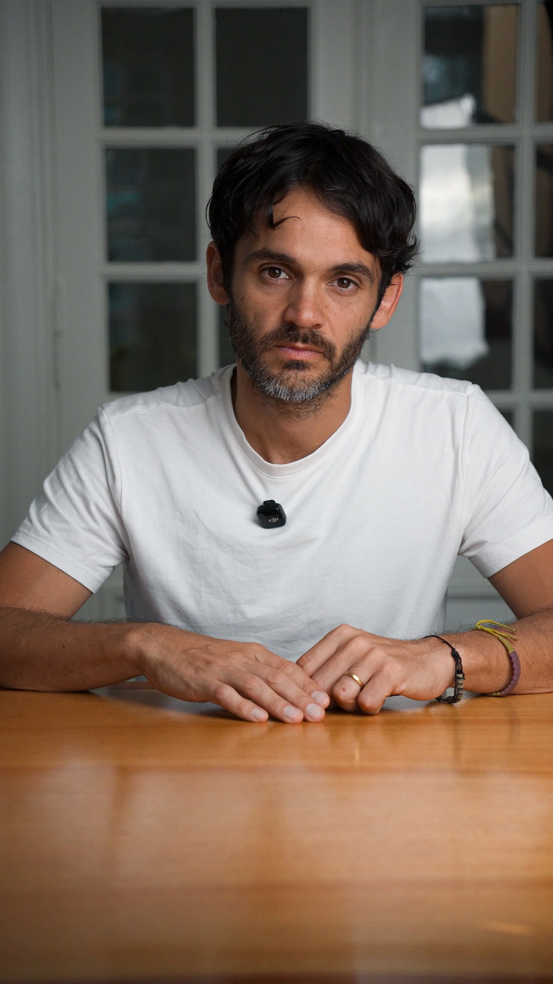 Hombre con barba y cabello oscuro, usando camiseta blanca, sentado en una mesa de madera, con fondo de ventanas de cristal.