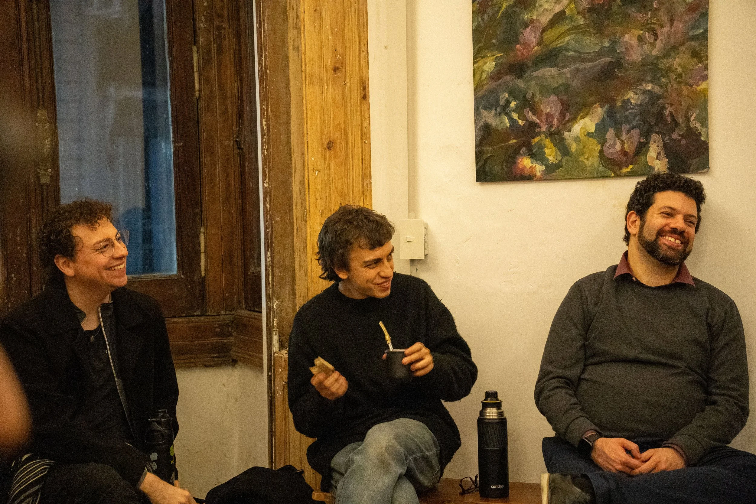 Tres personas están sentadas en una habitación, una sonríe, otra está tomando algo en una taza y la tercera está sonriendo. En la pared hay una pintura abstracta y una ventana de madera.