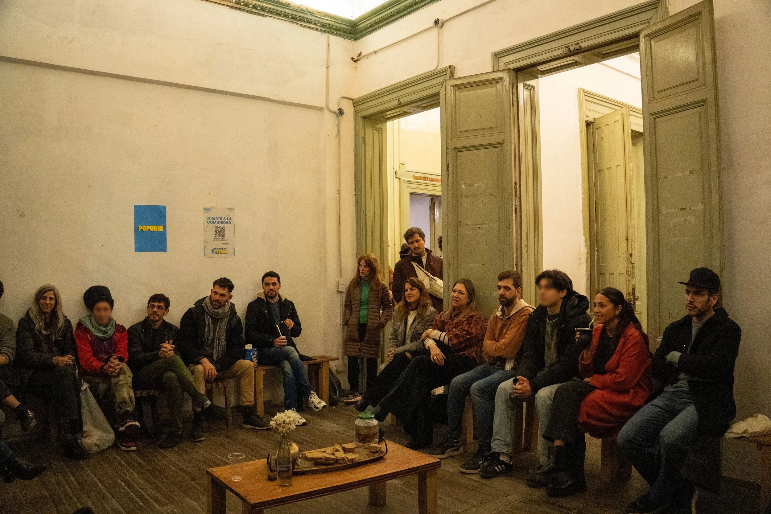 Grupo de personas sentadas y de pie en una sala con paredes blancas y puertas altas y abiertas, participando en una reunión o evento social.