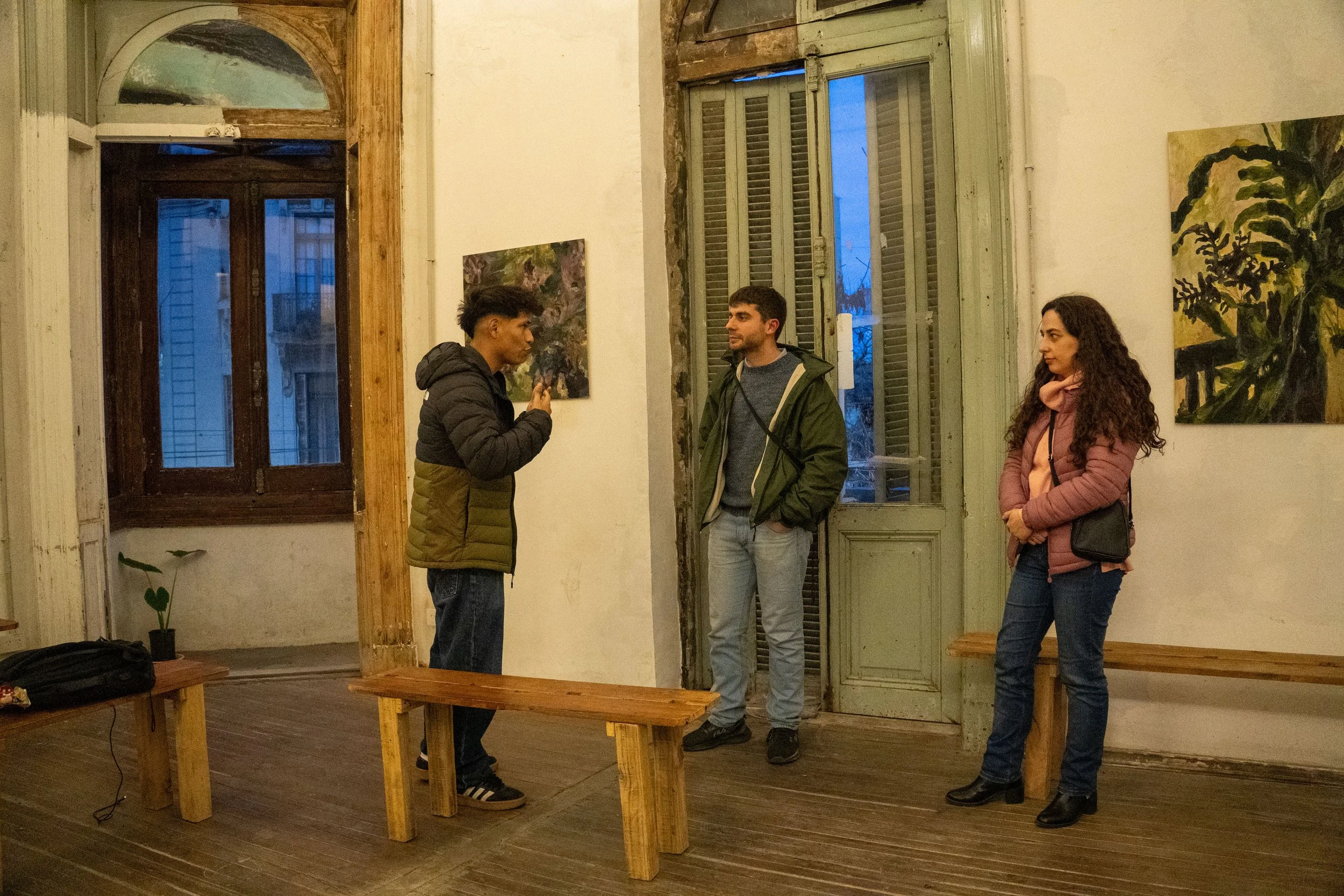 Tres personas conversando en una galería de arte con pinturas en las paredes, en un entorno con ventanas de madera y paredes blancas.