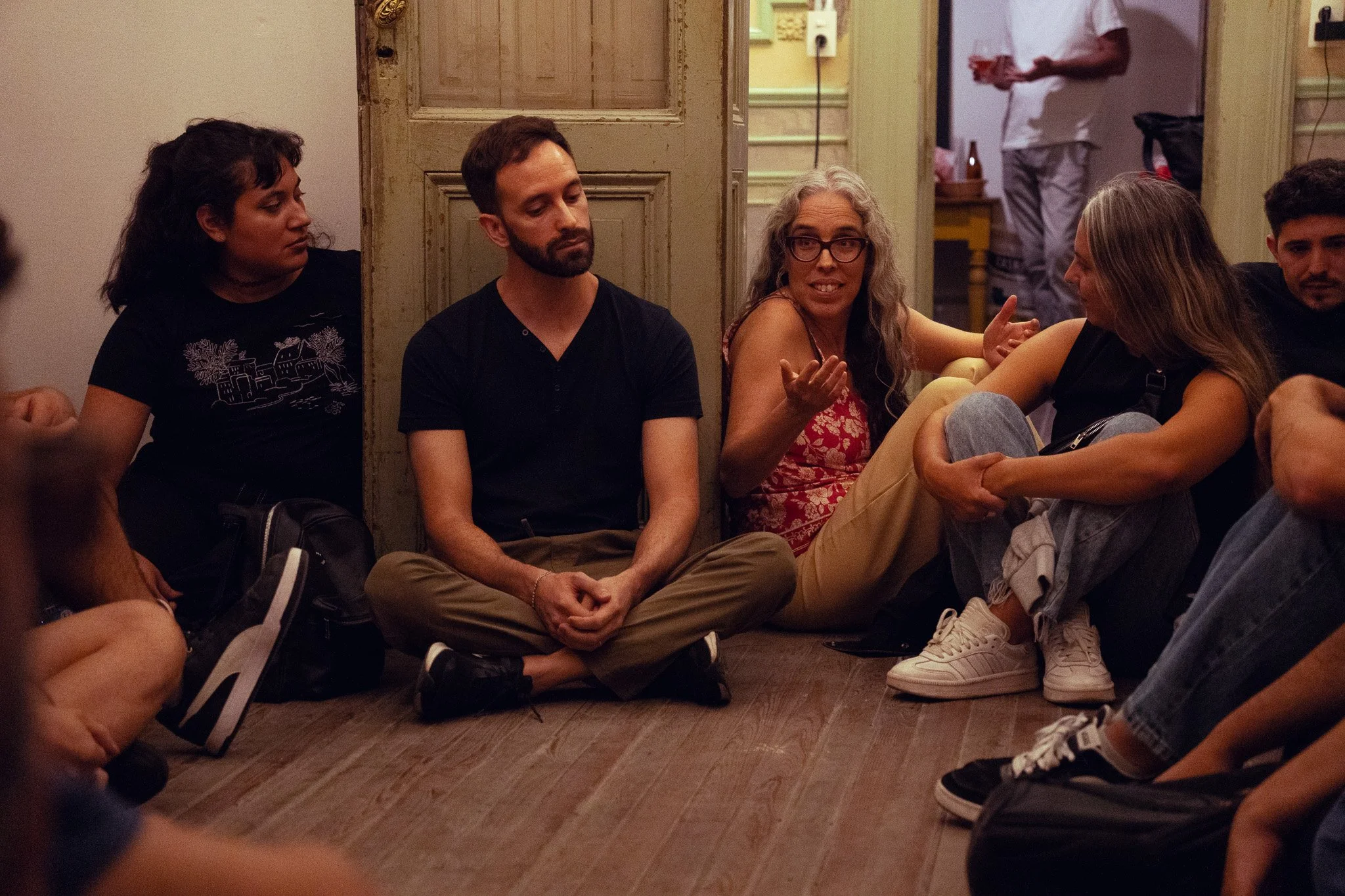 Grupo de personas sentadas en el suelo durante una conversación en una habitación con paredes de madera y puerta antigua, mientras una mujer con gafas habla y gesticula con las manos.